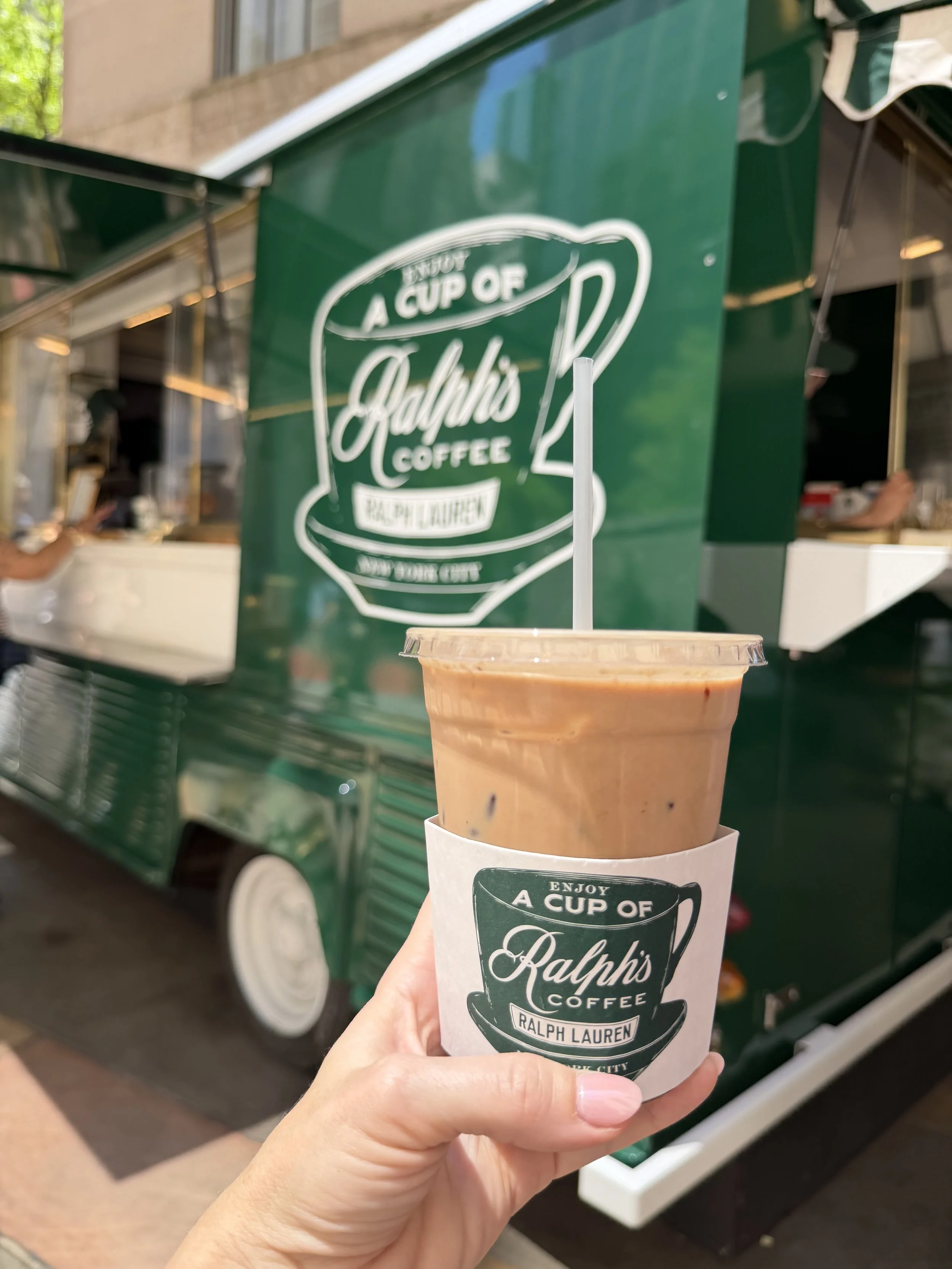 Cold coffee beverage in a clear plastic cup with a plastic lid and straw, in front of a green food truck with Ralph's Coffee branding.