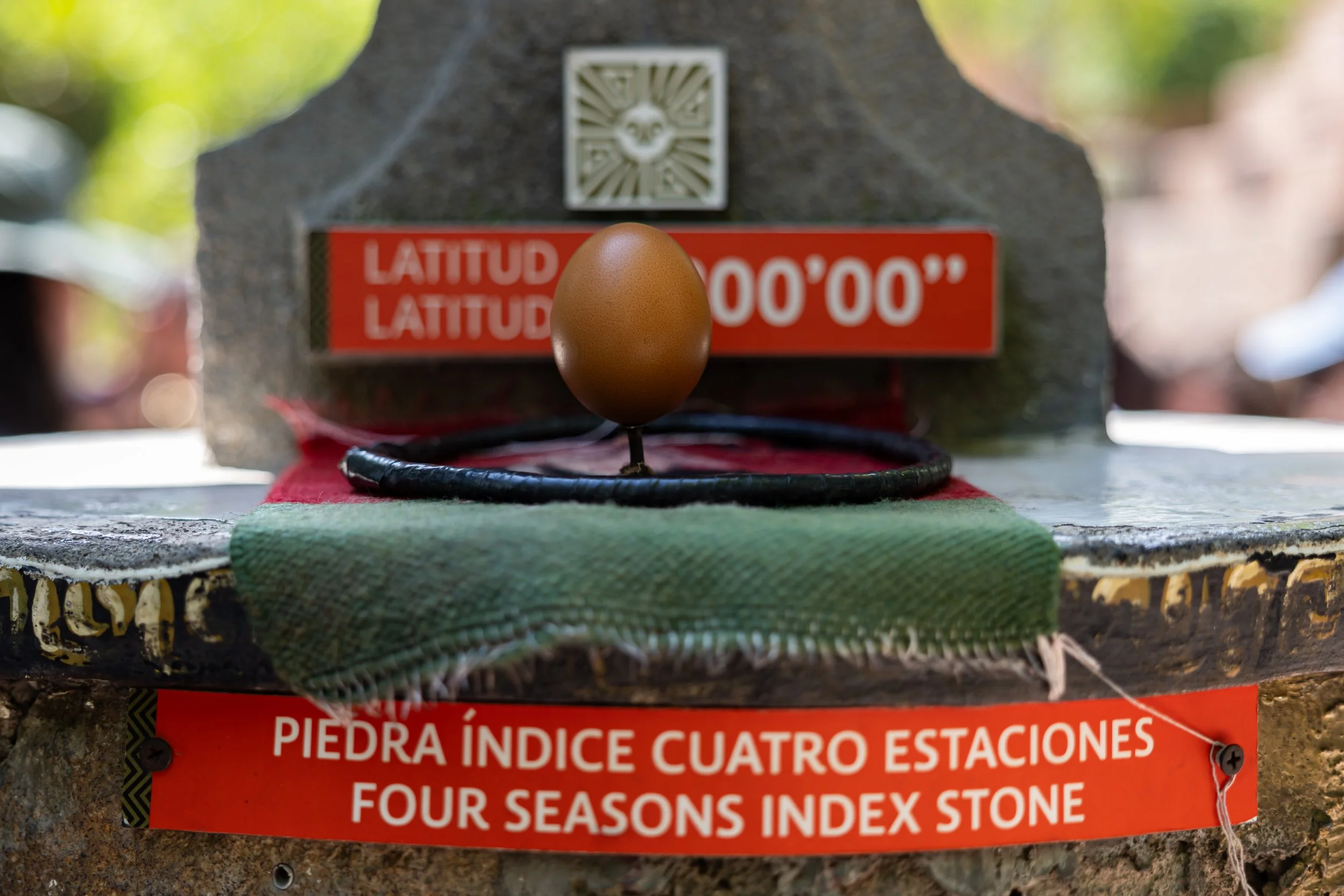 A stone marker with a metal emblem and a red sign that reads 'Four Seasons Index Stone' in Spanish and English, with a single brown egg balanced on a black wire in front of it.