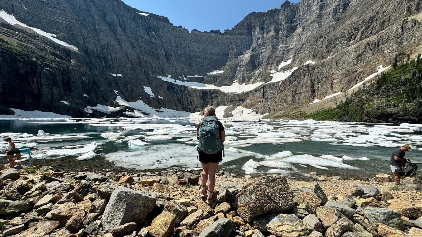 Morgan with a backpack standing on rocky shore in front of a glacier-fed lake with floating ice patches surrounded by tall, rugged mountains.