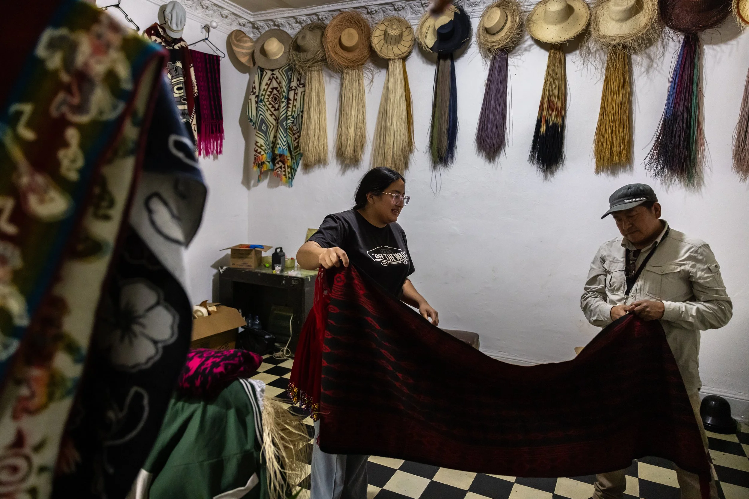 People sitting and standing in a room with hats hanging on the wall, and colorful textiles and souvenirs on display.