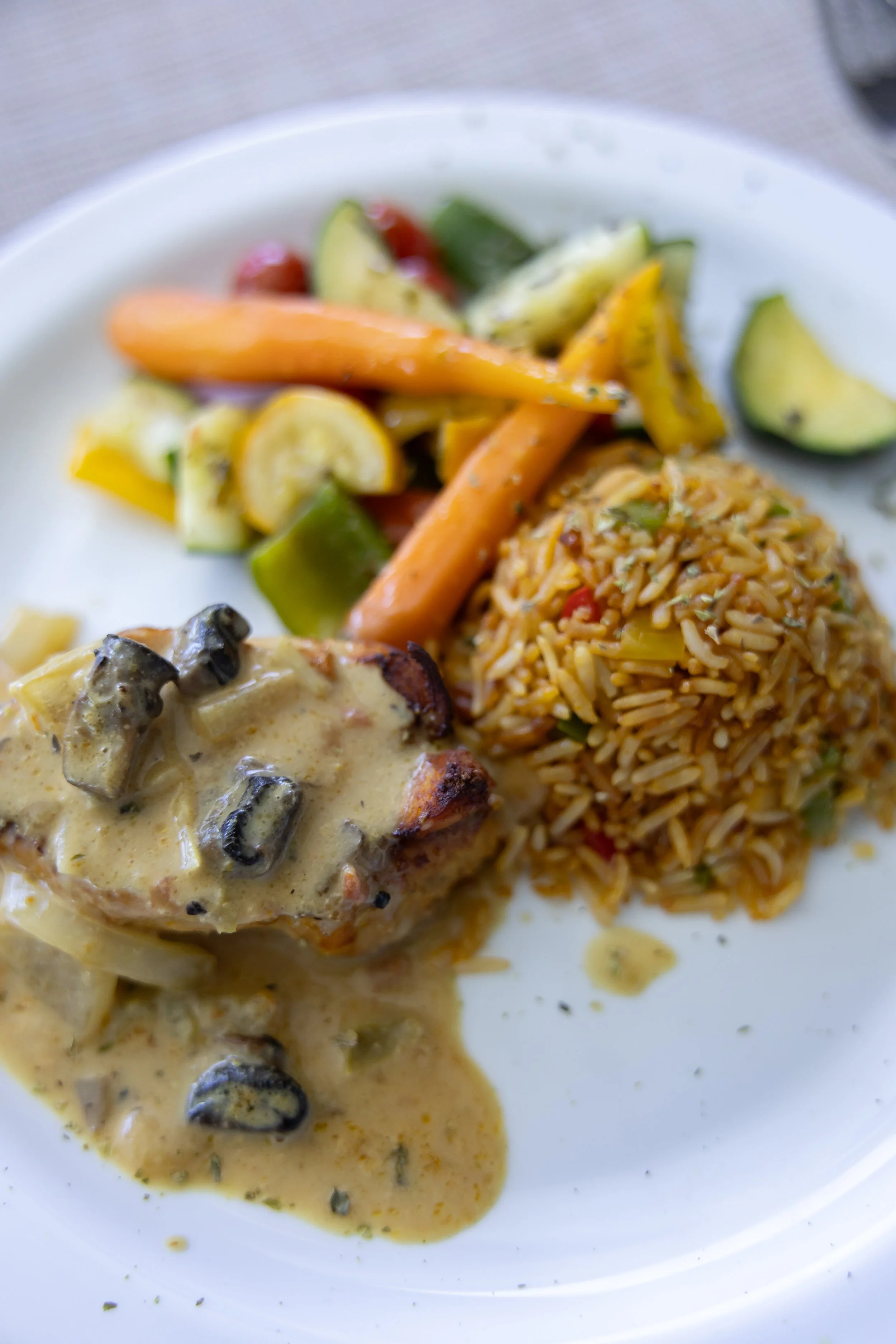 Plate of food with a piece of chicken covered in creamy mushroom sauce, mixed rice, and assorted vegetables including carrots, zucchini, and cherry tomatoes.