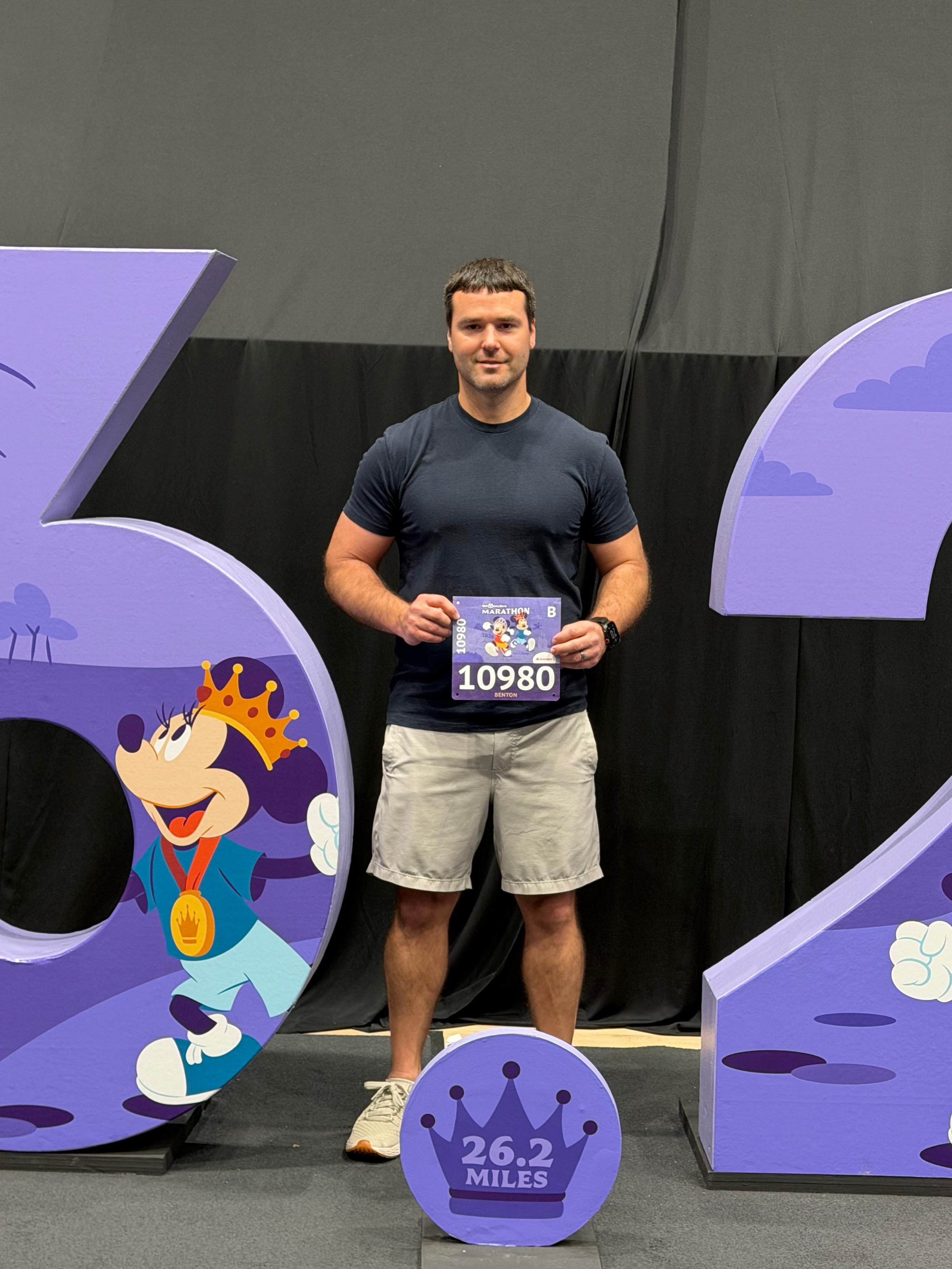Man with race bib, at RunDisney event, holding bib, standing at the expo, in front of 26.2 mile sign