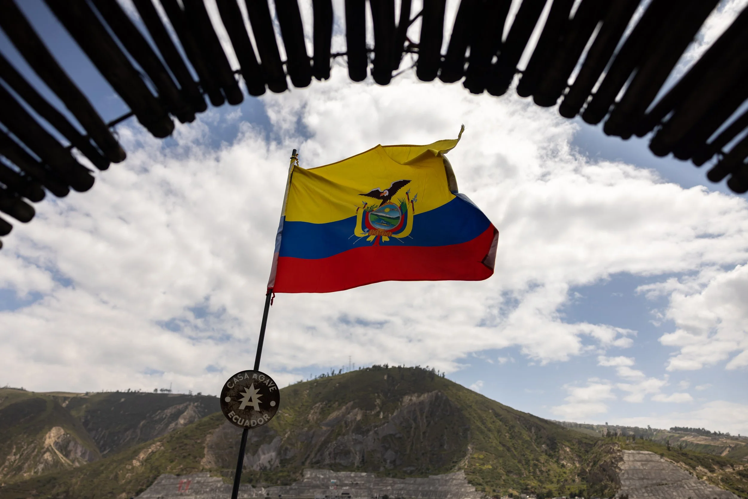 Ecuador flag flying against cloudy sky with mountains and hillside in the background.