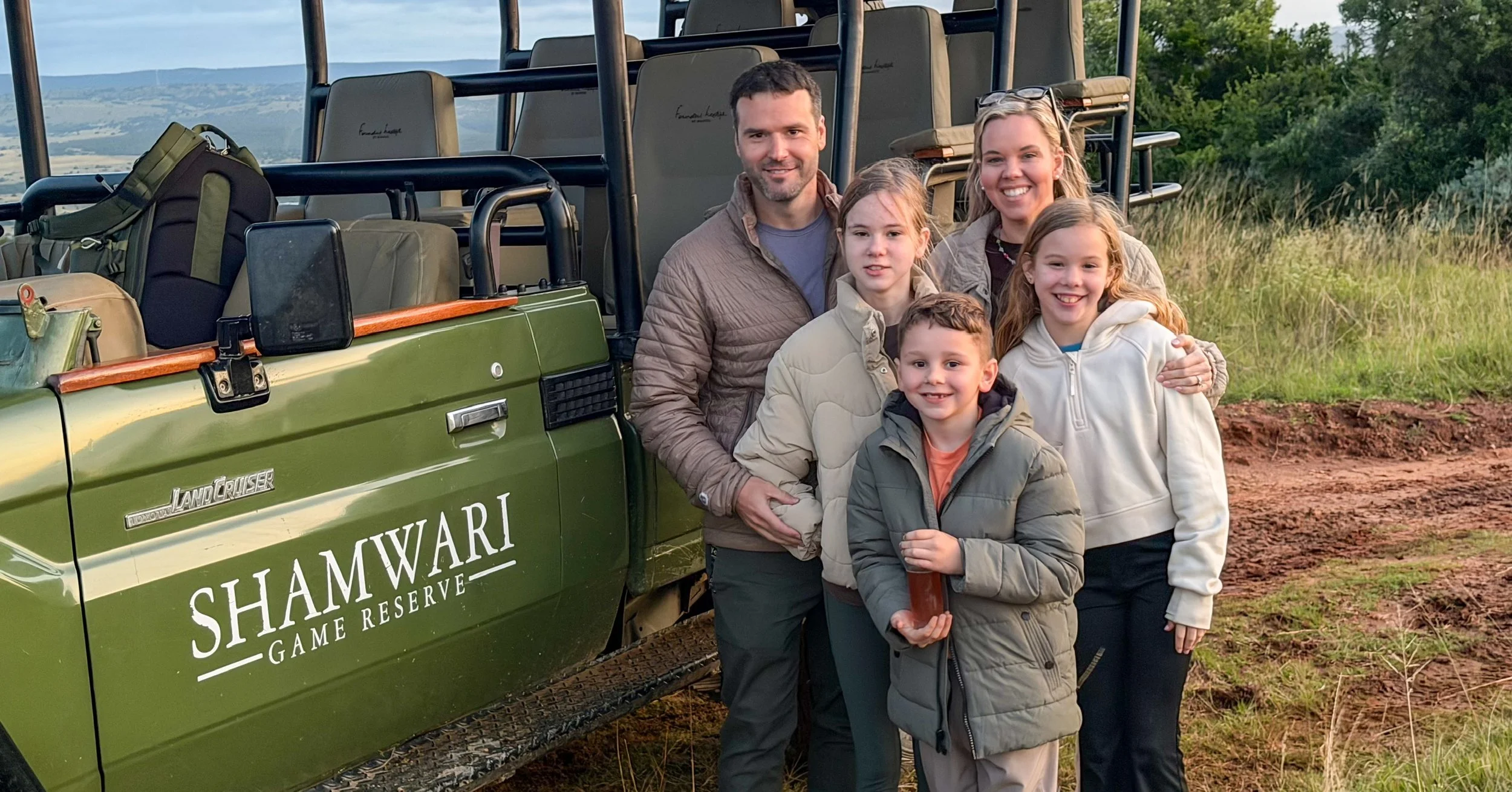 Sanders Family posing outdoors next to a green Land Cruiser with 'Shamwari Game Reserve' written on the side. They are on a dirt path with trees and rolling hills in the background, during daytime.