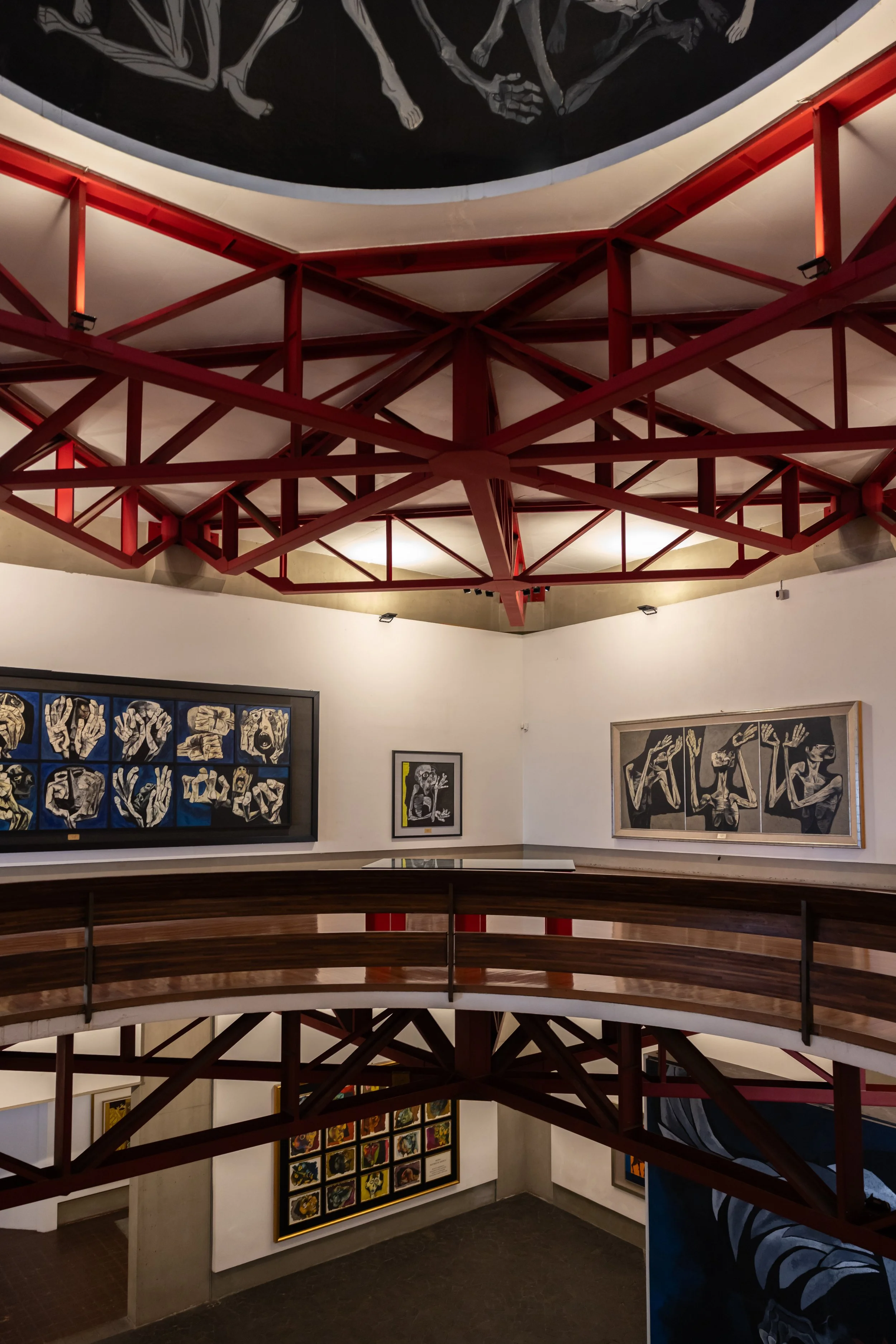 Photo of multiple artworks displayed on gallery walls, viewed from a balcony with a red steel framework overhead, featuring paintings with space and animal themes.