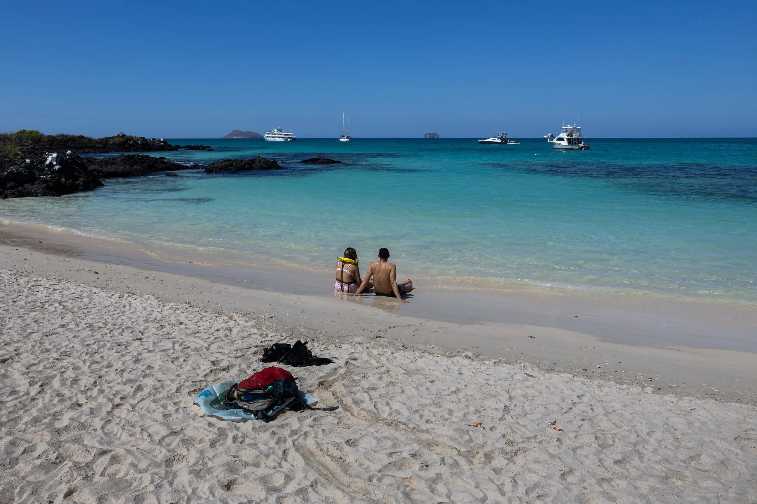 A sandy beach with two people sitting on the shore facing the ocean. Several boats are anchored offshore, with a rocky island and small landmass in the background under a clear blue sky.