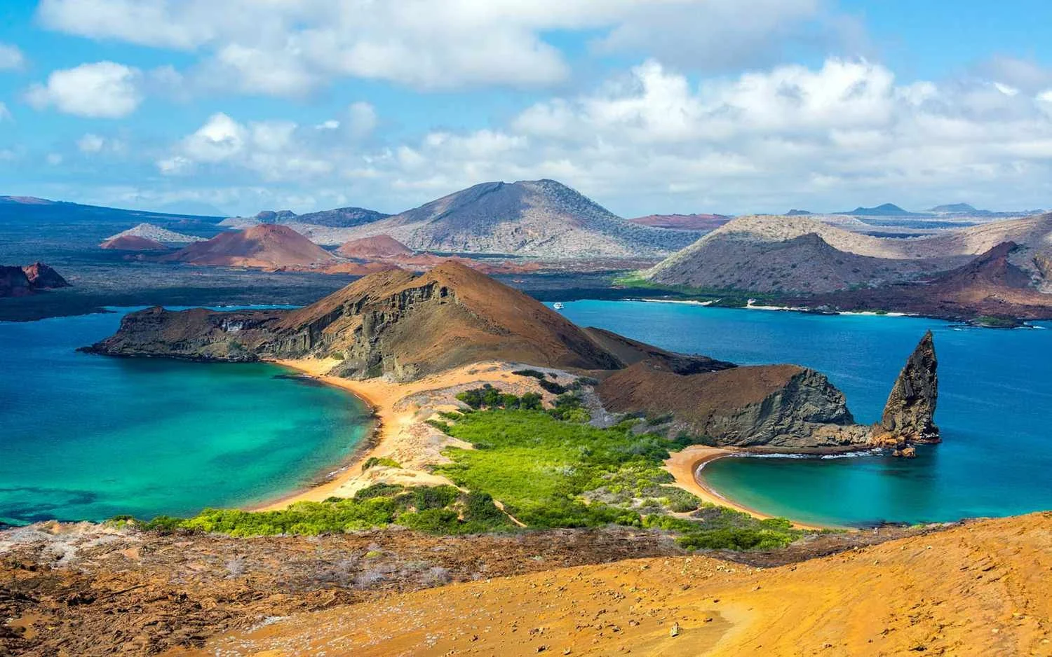 A scenic view of a volcanic island with two turquoise bays, green vegetation, rocky formations, and distant mountains under a partly cloudy sky.