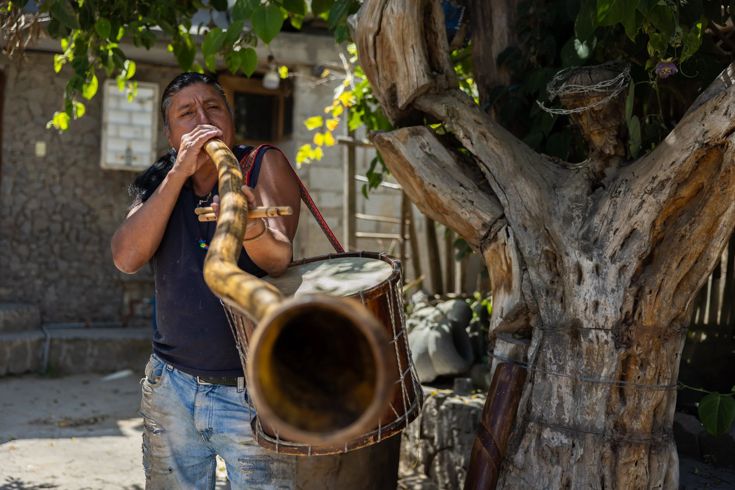 A woman is playing a traditional wind instrument outdoors near a large tree with thick, textured bark and green leaves. She is wearing a dark sleeveless top and light jeans, and has long dark hair tied back. The background shows a rustic stone buildi