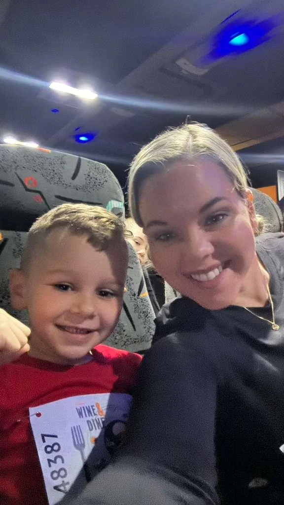 A woman and a young boy sitting on a bus, both smiling at the camera. The boy is wearing a red shirt with a name tag. The bus seats are gray with a speckled pattern, and the ceiling has blue and white lights.