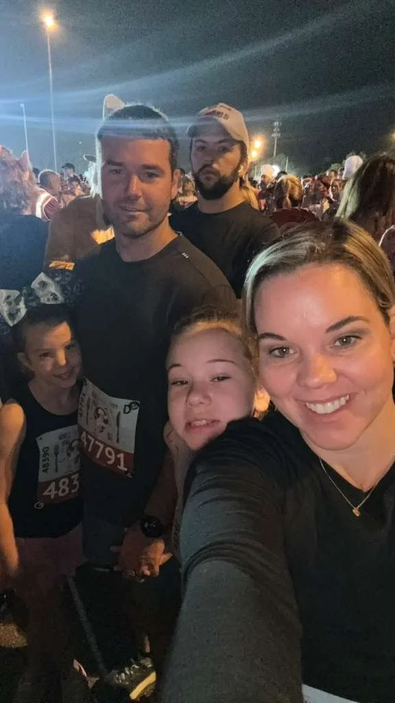 A group of five people, including children and adults, taking a selfie at a nighttime outdoor event with many other people in the background. Two children are wearing race bibs, indicating a running event.