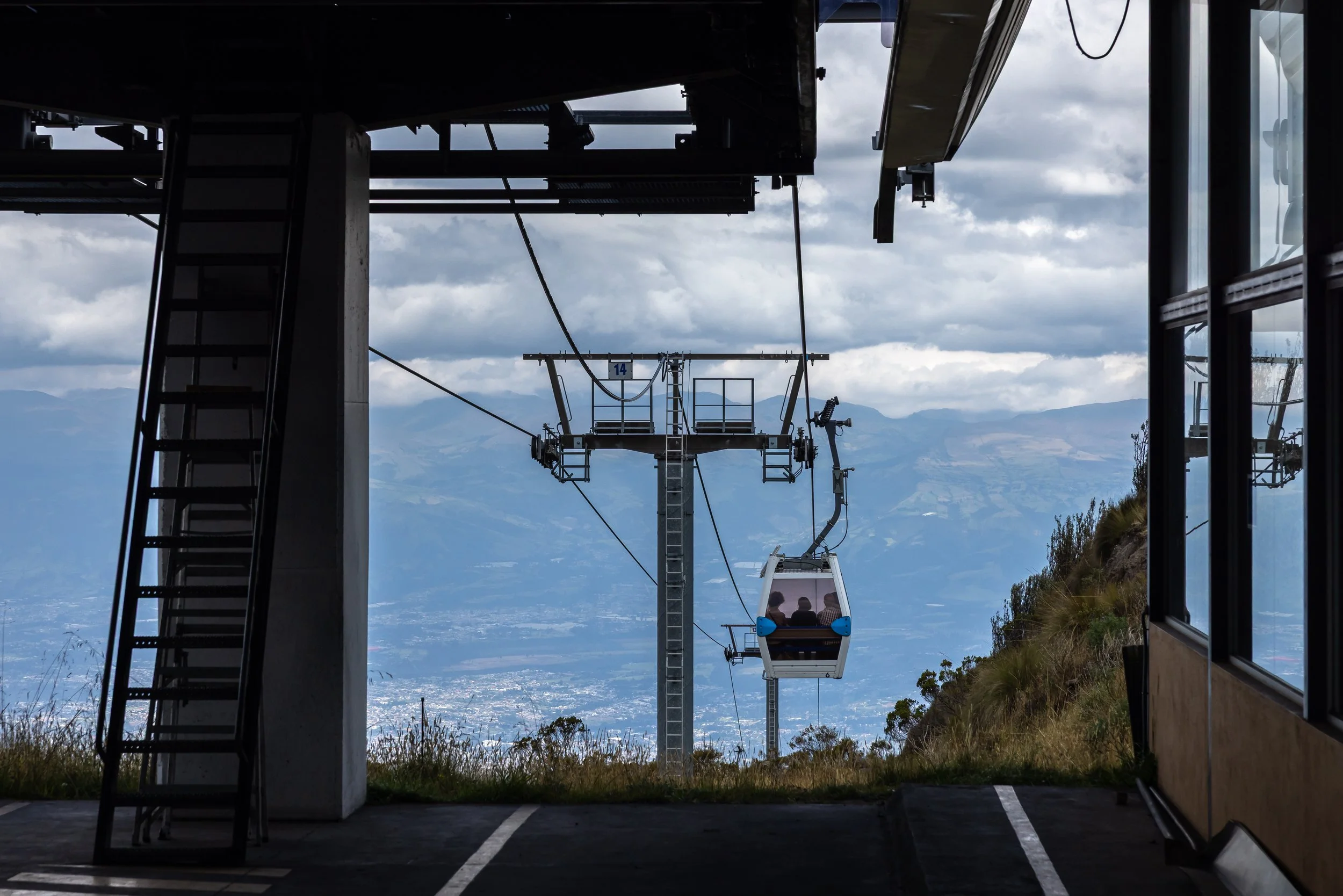 Cable car ascending a mountain with views of the valley and mountains in the background. The photo is taken from inside a structure with a ladder on the left and framed windows on the right.