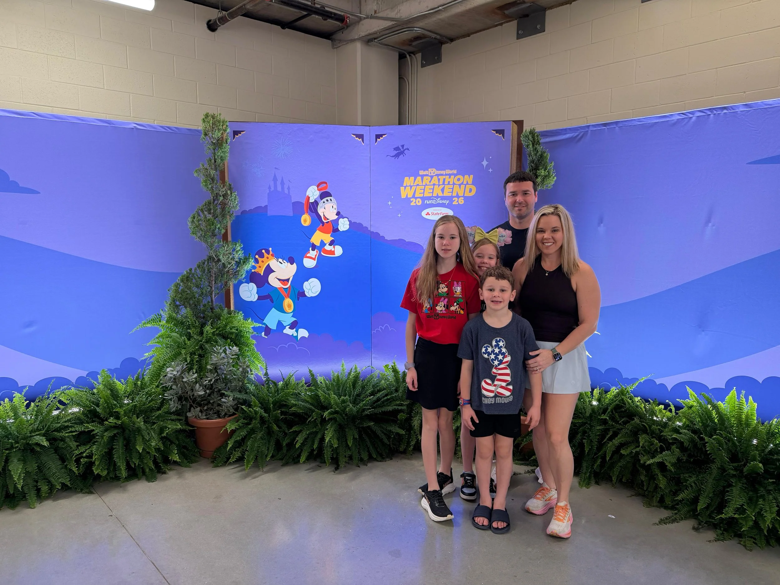 A group of five people, including two women and three children, standing in front of a Disney-themed backdrop with characters Mickey Mouse and Minnie Mouse and the text "Marathon Weekend 2026." They are surrounded by potted ferns and greenery.