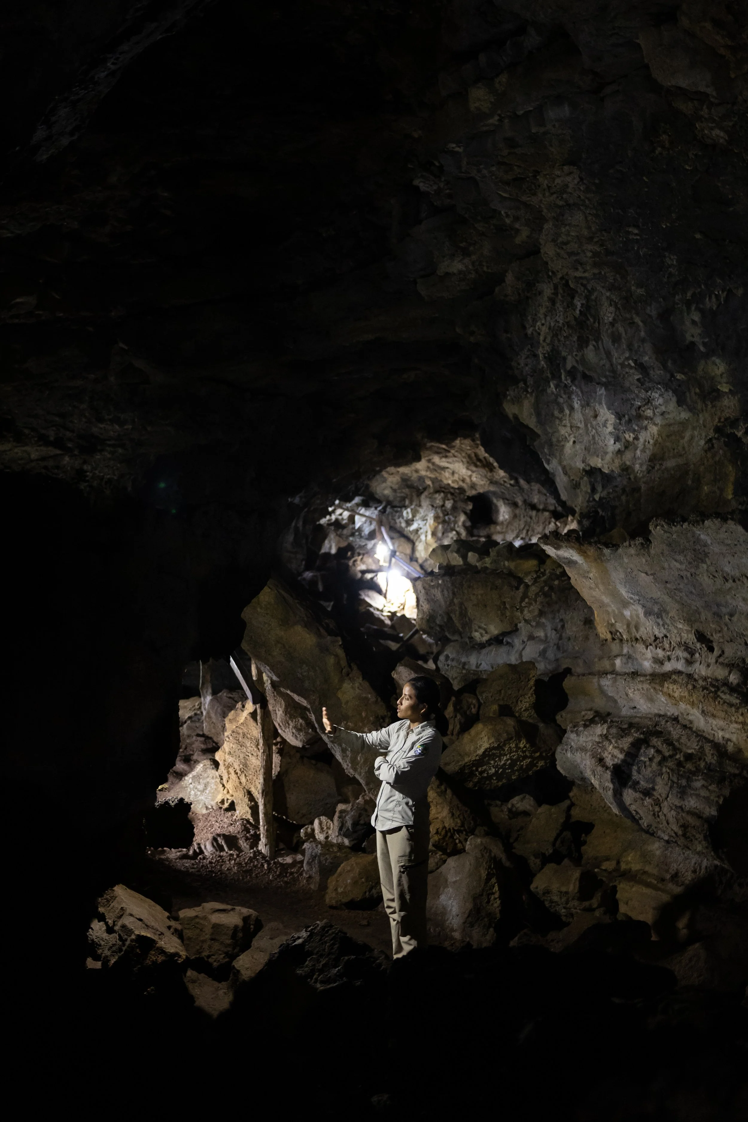 A person wearing a gray jacket and light pants standing inside a dark cave, reaching out with their right hand and looking at the rocky ceiling.