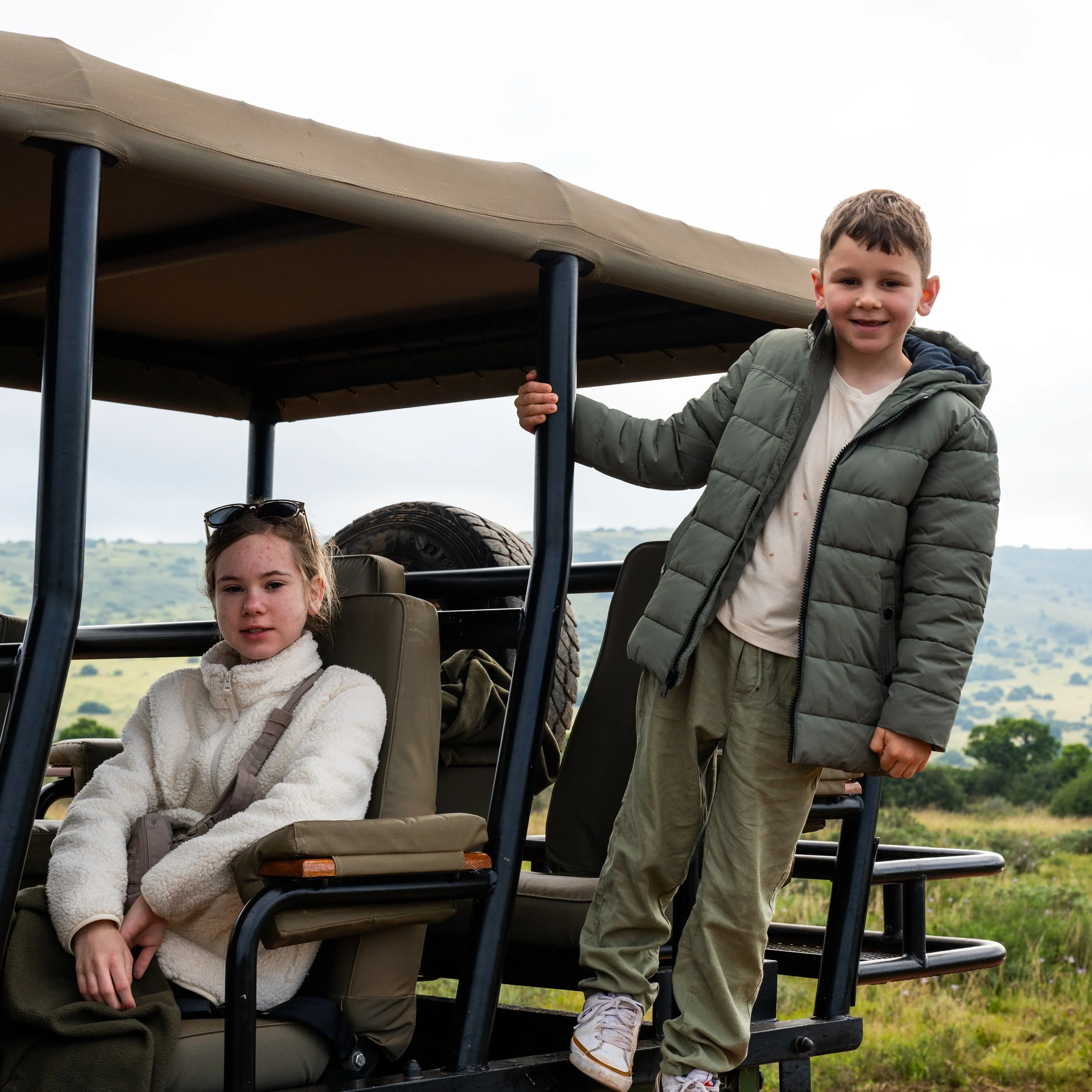 Rae & Jed riding in a safari-style vehicle with open sides, overlooking a rural landscape with hills and trees in the background.
