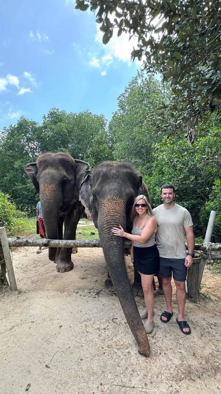 Morgan & Benton stands with two elephants in a lush outdoor setting with green trees and a bright blue sky.