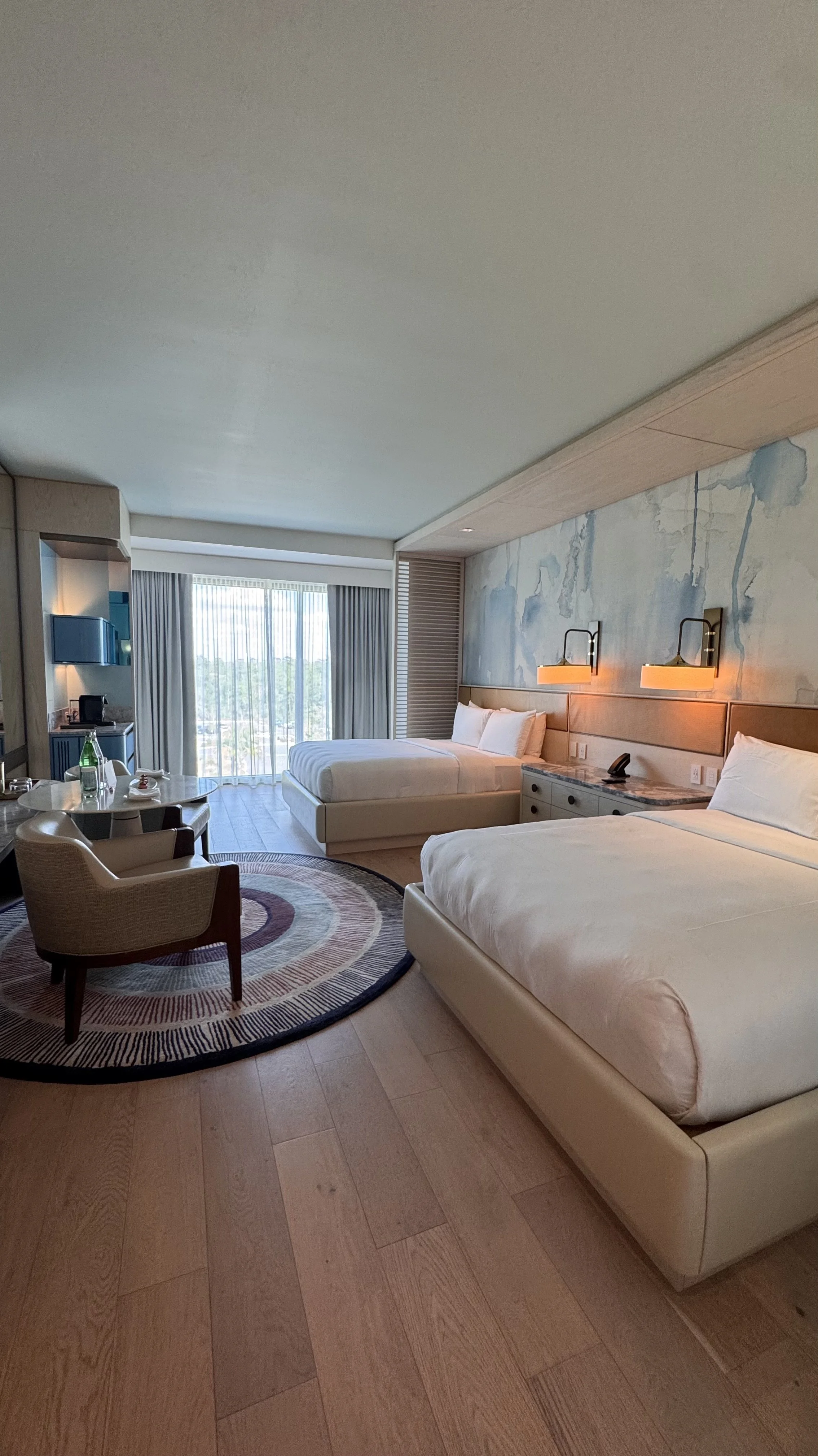 Hotel room with two beds, a sitting area, and a view of a city through large windows.