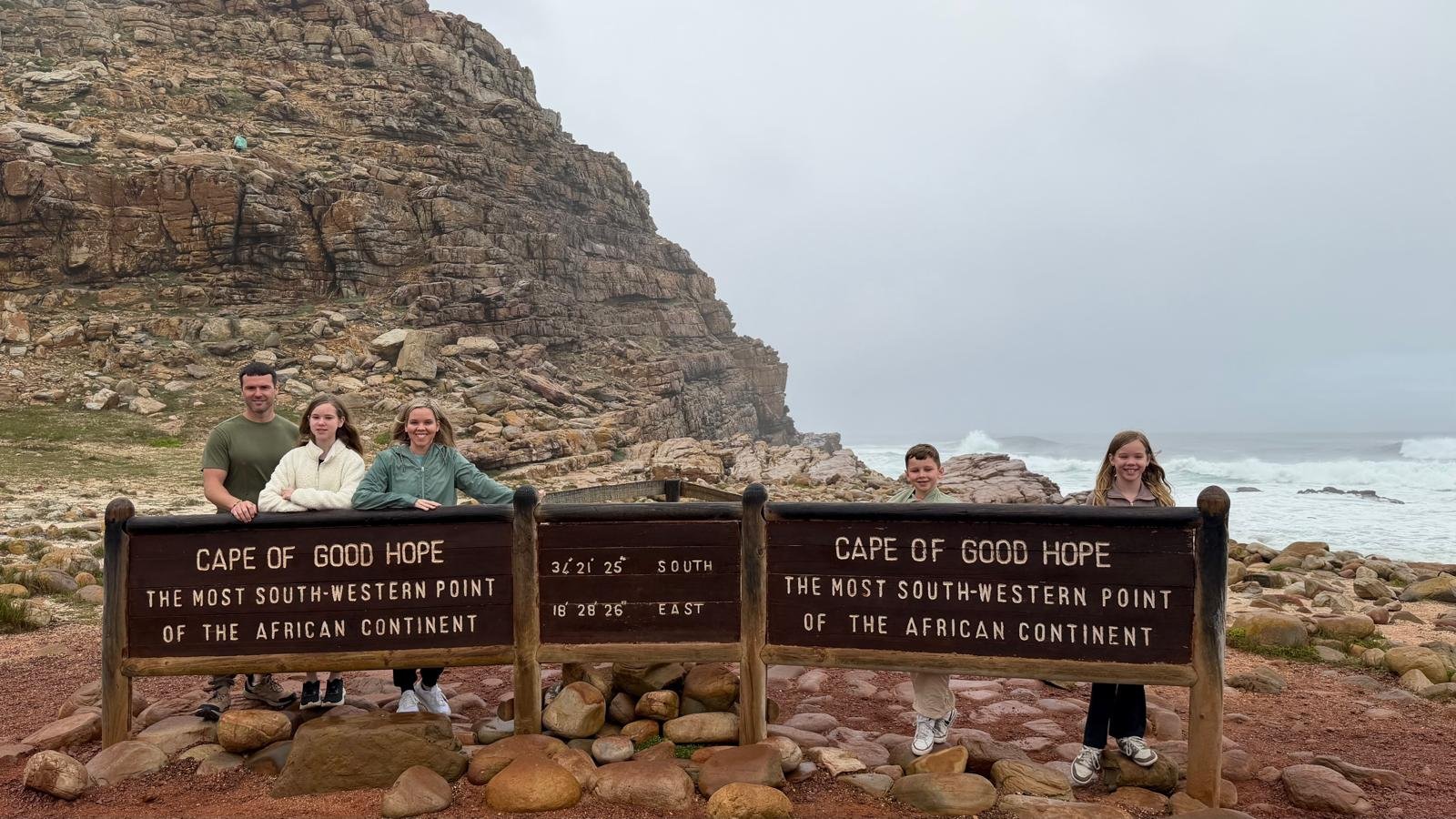 Sanders Family stand behind signs at the Cape of Good Hope, with rocky cliffs and the ocean in the background. The signs indicate it's the southwesternmost point of the African continent.