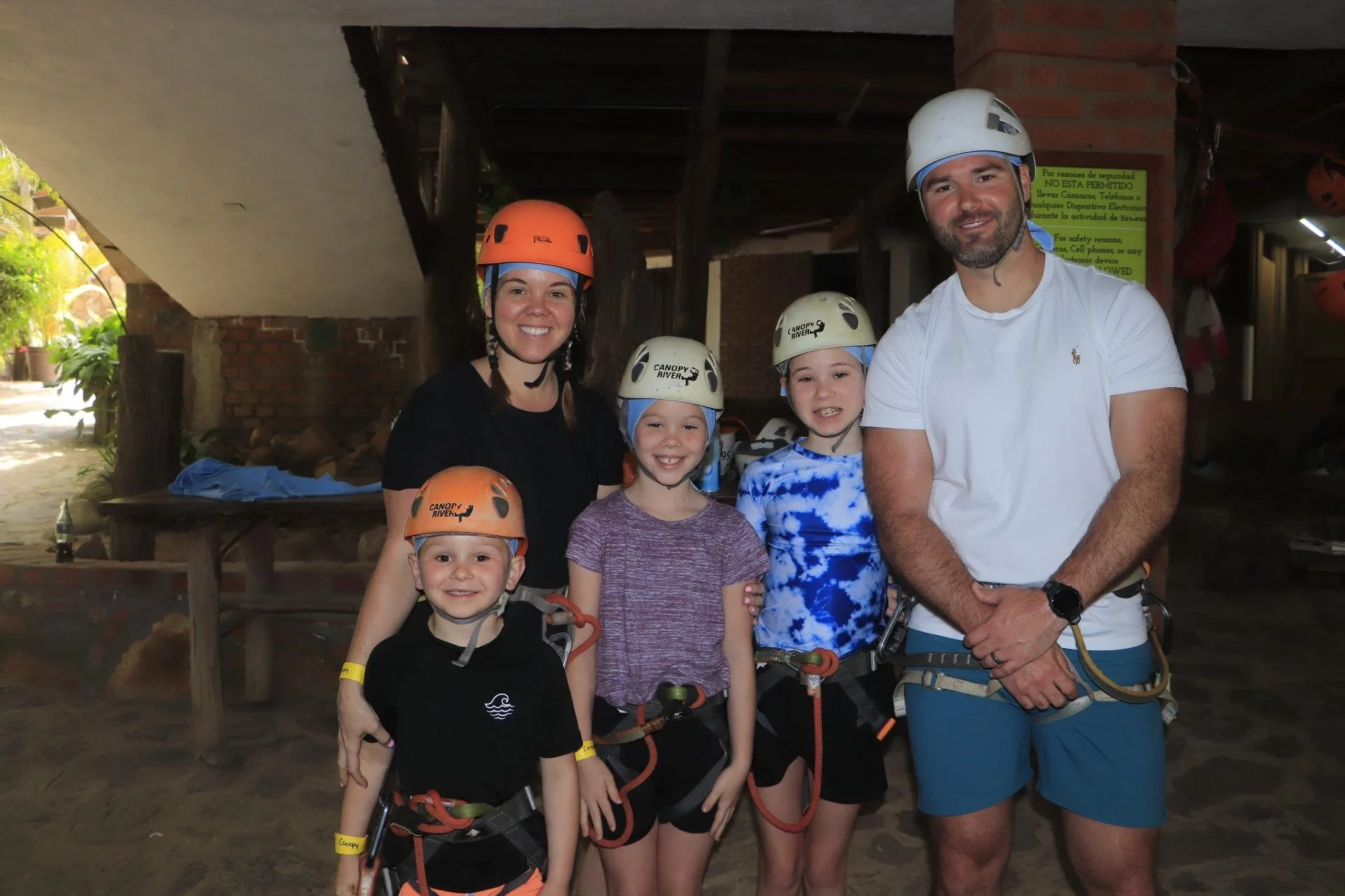 Sanders Family, wearing helmets and harnesses, standing together indoors with a brick and wood structure in the background. The group is smiling at the camera, likely preparing for an outdoor adventure activity.