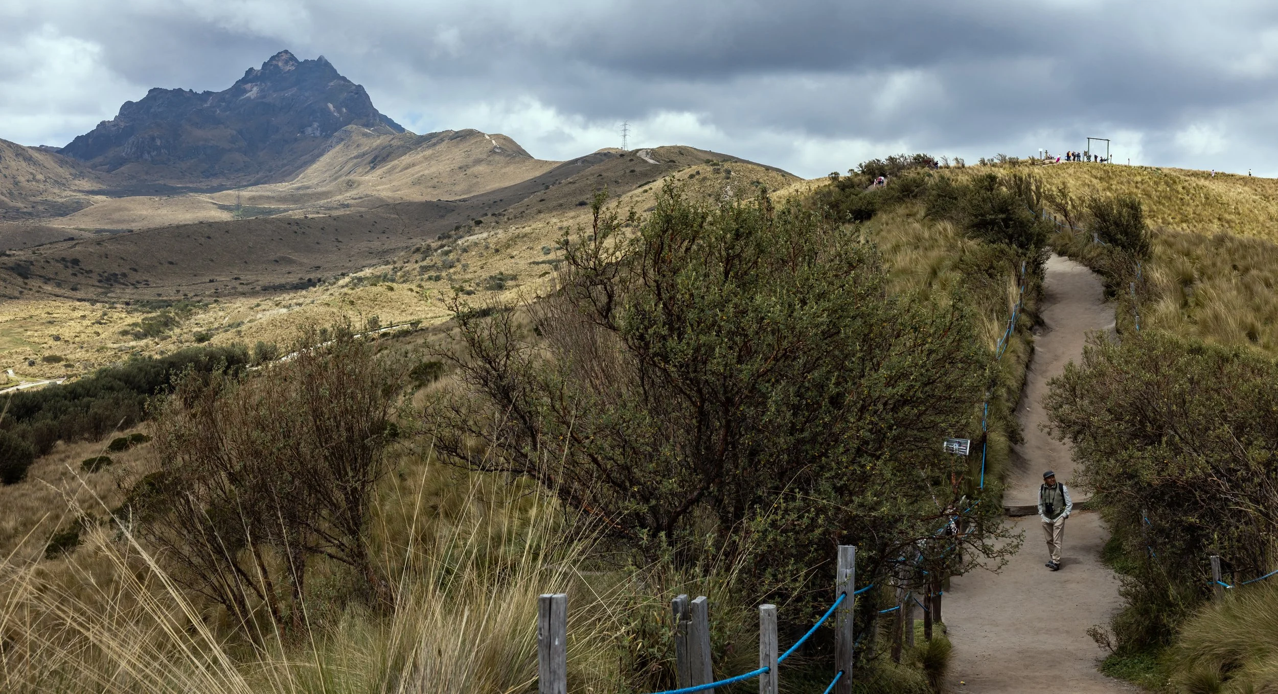 A hiker walking down a trail in a mountainous landscape with hills and a mountain peak in the background, under a cloudy sky.