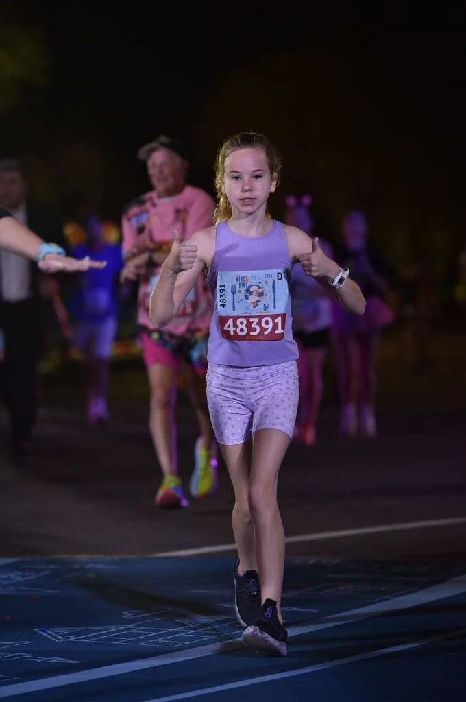 A young girl running in a race during nighttime or evening, wearing a purple tank top and patterned shorts, with a race bib number 48391, and surrounded by other runners and spectators.