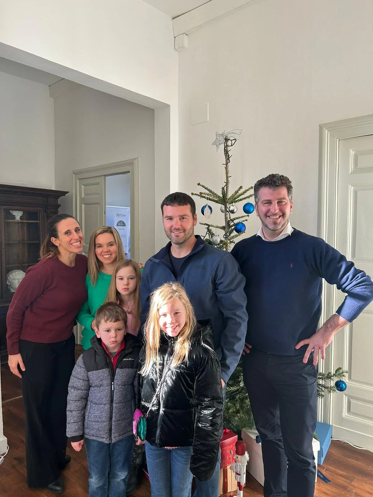 A group of seven people, including adults and children, standing in front of a decorated Christmas tree with blue and silver ornaments in a bright room.