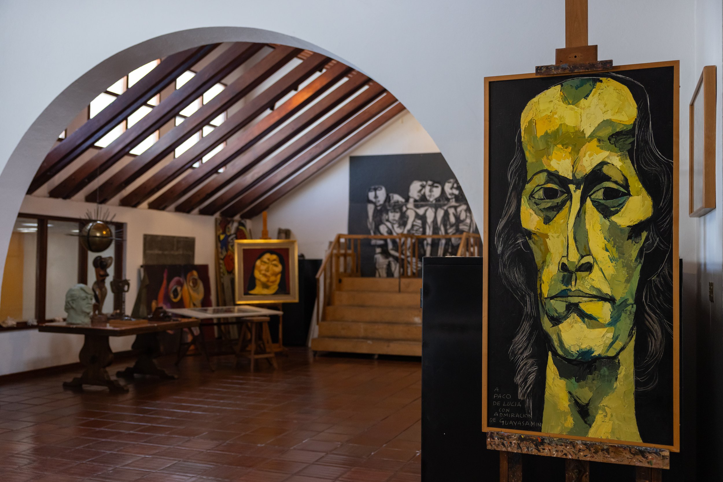 Art gallery interior with paintings, sculptures, and wooden staircase under a sloped ceiling with wooden beams.
