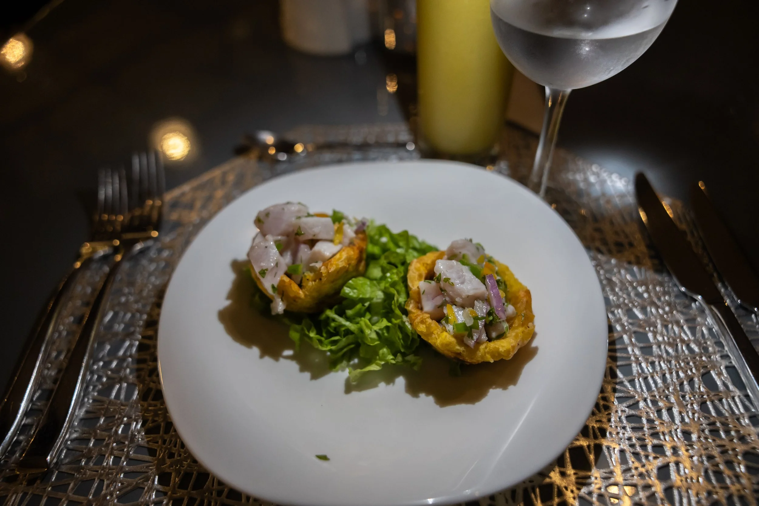 Two fried shells topped with shrimp and diced vegetables on a bed of lettuce, served on a white plate with a glass of water and green juice in the background, on a decorative gold and glass table.