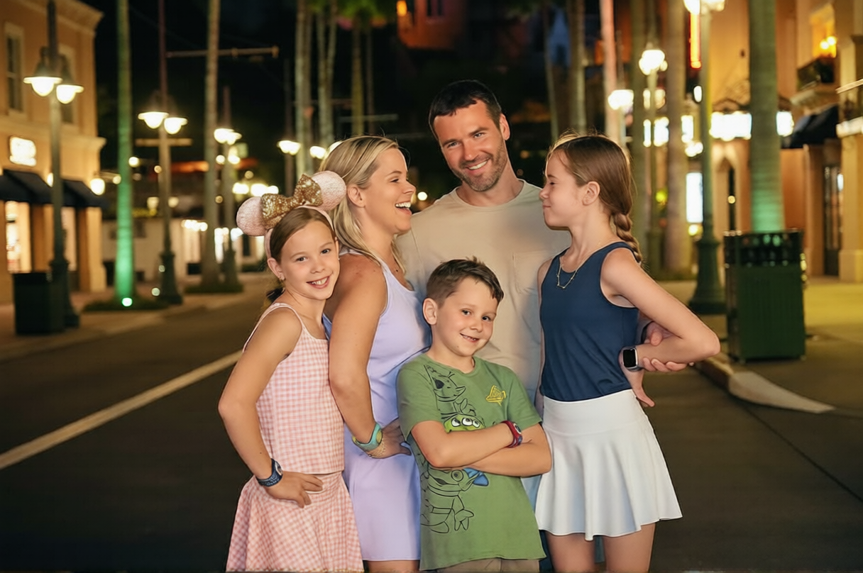 A family of five posing together on a city street at night, with illuminated buildings and streetlights in the background. The family members are smiling and appear happy.