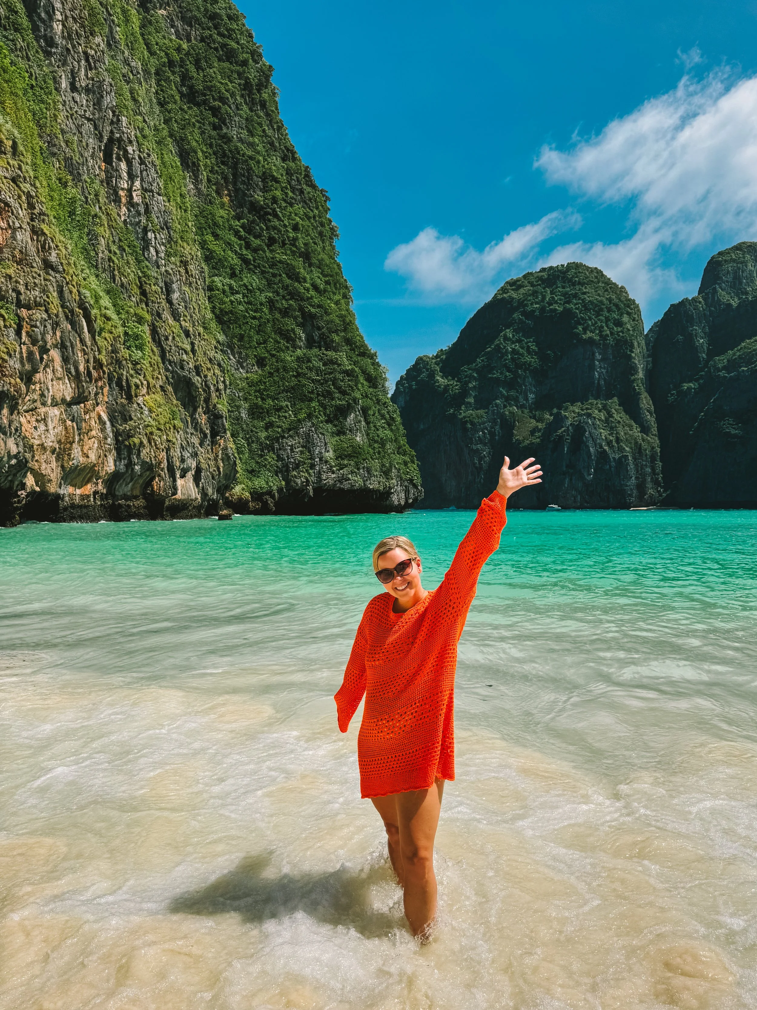 Morgan in sunglasses and an orange dress posing and smiling at the beach with green cliffs and blue sky in the background.