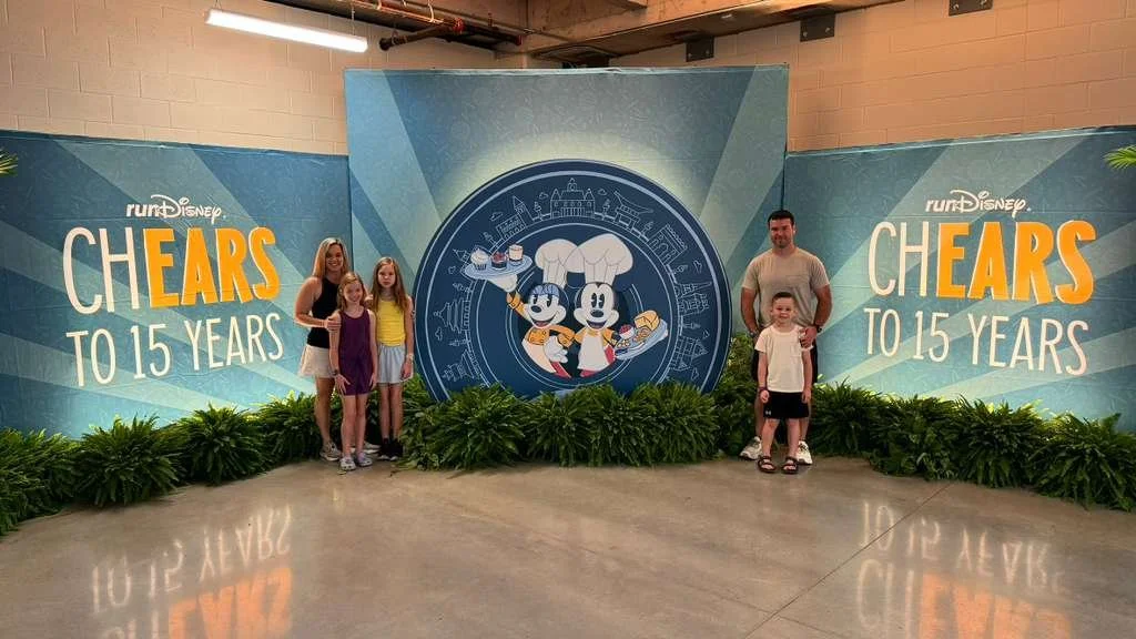 Family photo in front of a promotional backdrop for RunDisney's 'Cheers to 15 Years' event with three children, a woman, and a man, all standing on a polished floor with green plants at the base of the backdrop.