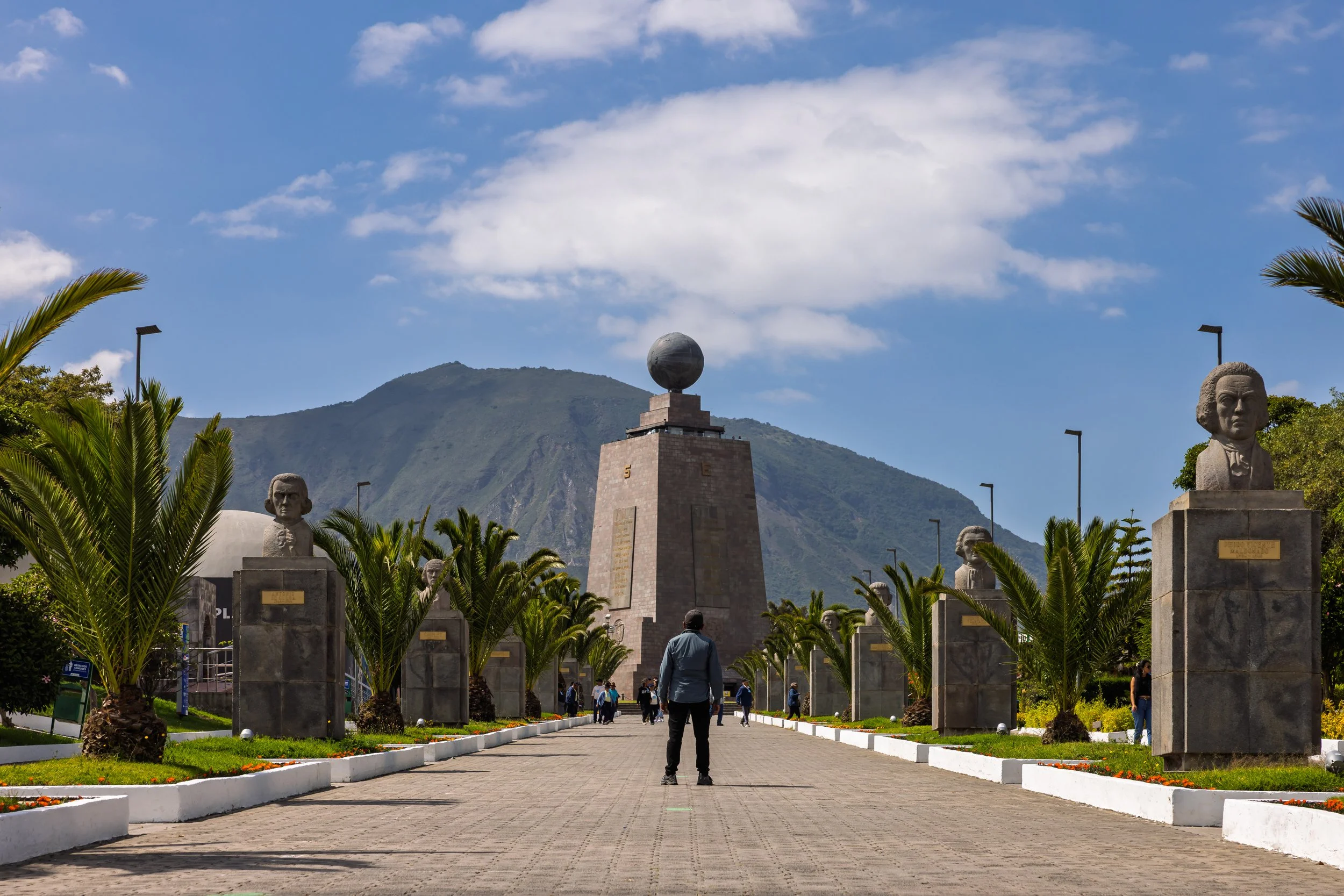 A memorial park with bust statues on pedestals lining a paved walkway, surrounded by greenery and tall streetlights, with a mountain in the background and blue sky with clouds above.