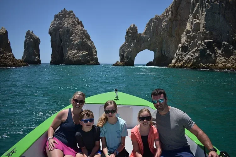 Sanders Family sitting on a green boat, sailing in the ocean near rocky cliffs and natural arches, under a clear blue sky.