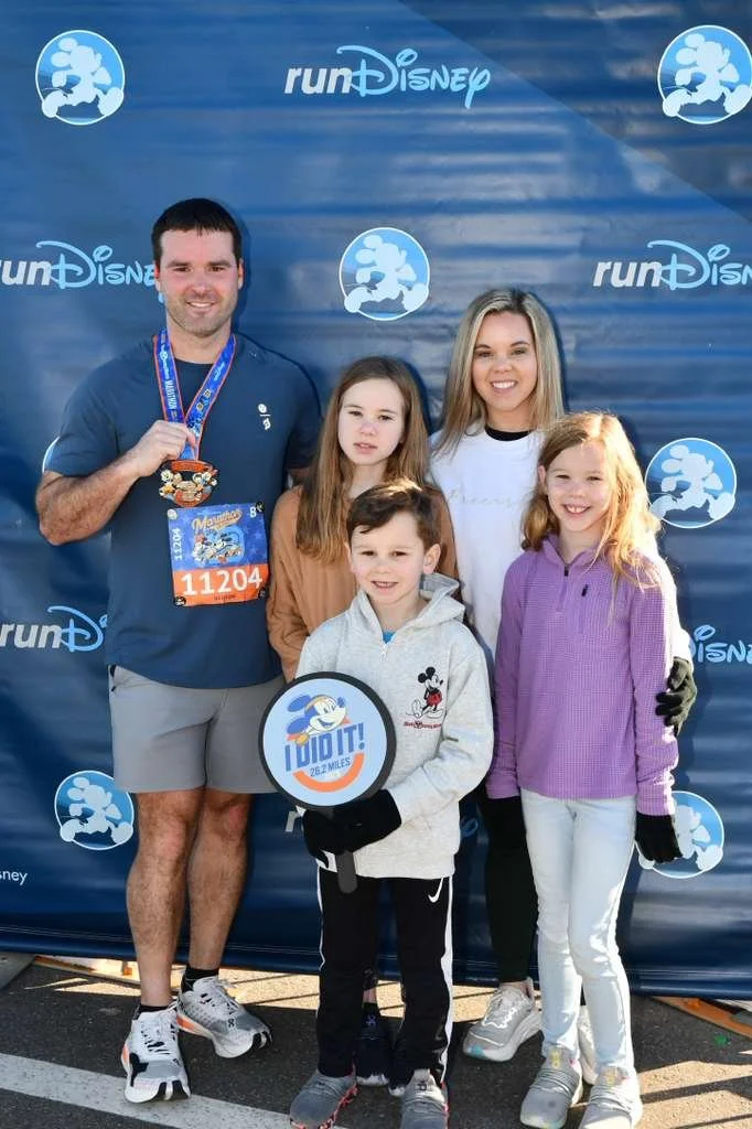 A family of five posing in front of a blue backdrop with 'runDisney' branding. The man is wearing a finisher medal and a race bib, holding it in his hand. The children are dressed casually, and one is holding a circular sign with the Disney mascot an