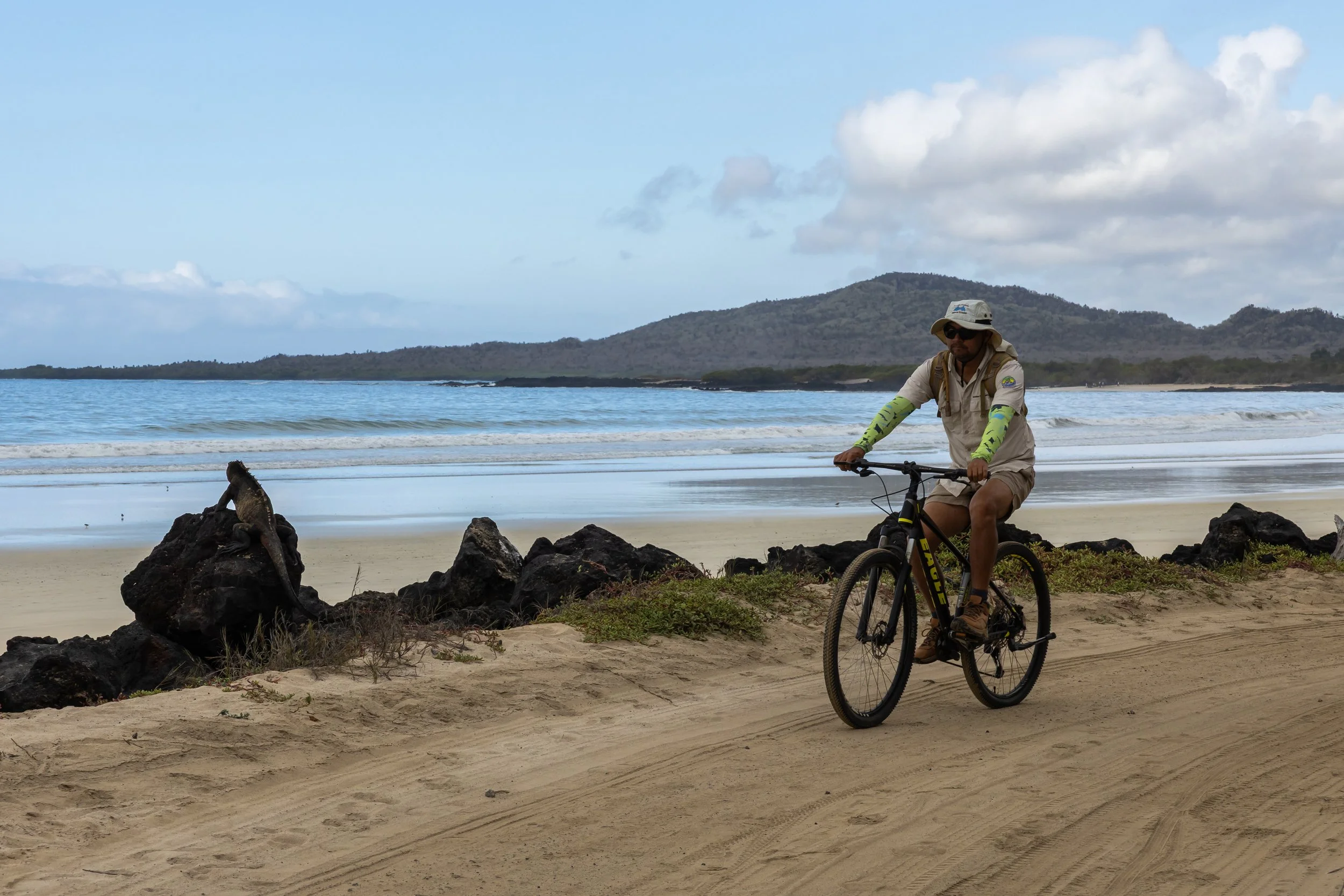 A man riding a bicycle on a beach near rocks with the ocean and mountains in the background, wearing sunglasses, a wide-brim hat, and outdoor clothing.