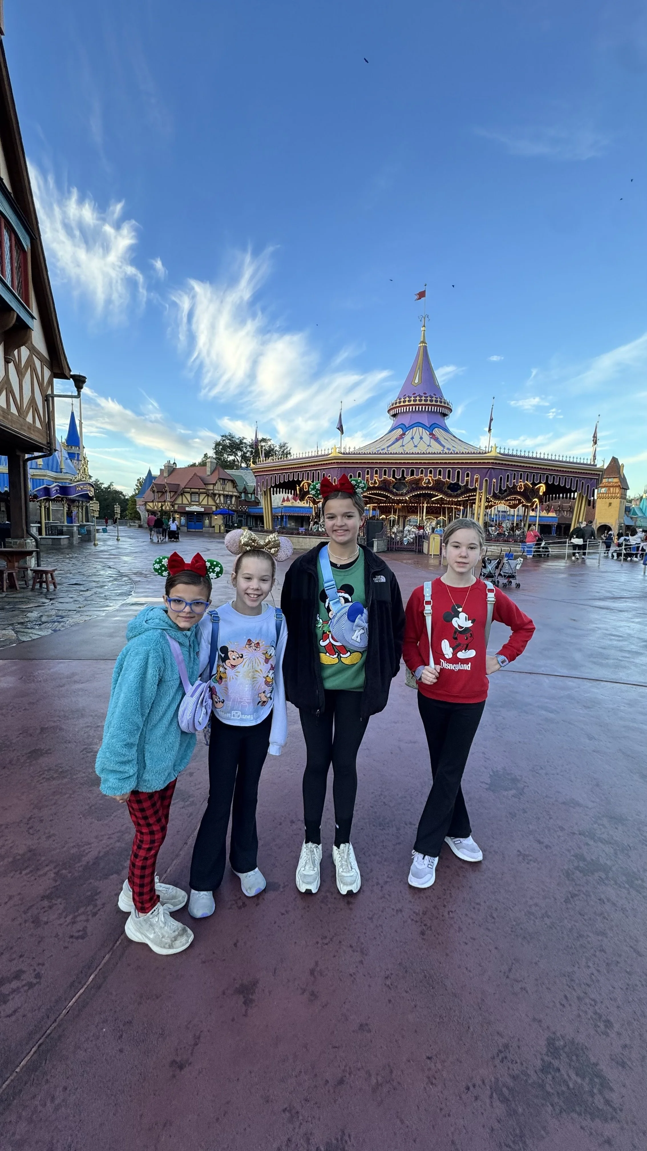 Sanders Family standing together at Disneyland with the Disneyland park in the background, featuring a carousel and colorful buildings, on a slightly cloudy day.