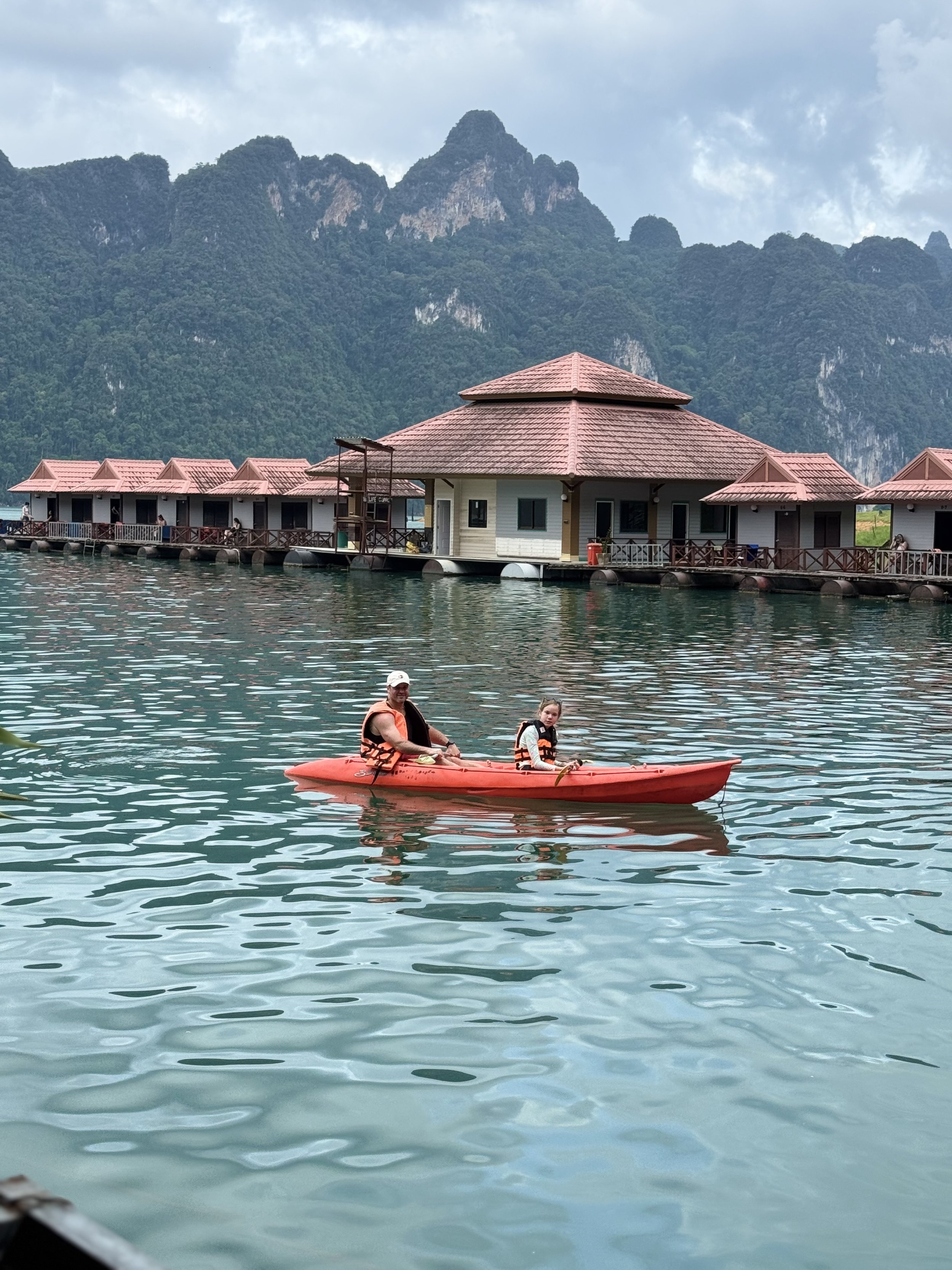 Father and daughter kayaking at Khao Sok National Park in front of floating houses on the lake in the summer time with the jungle in the background.