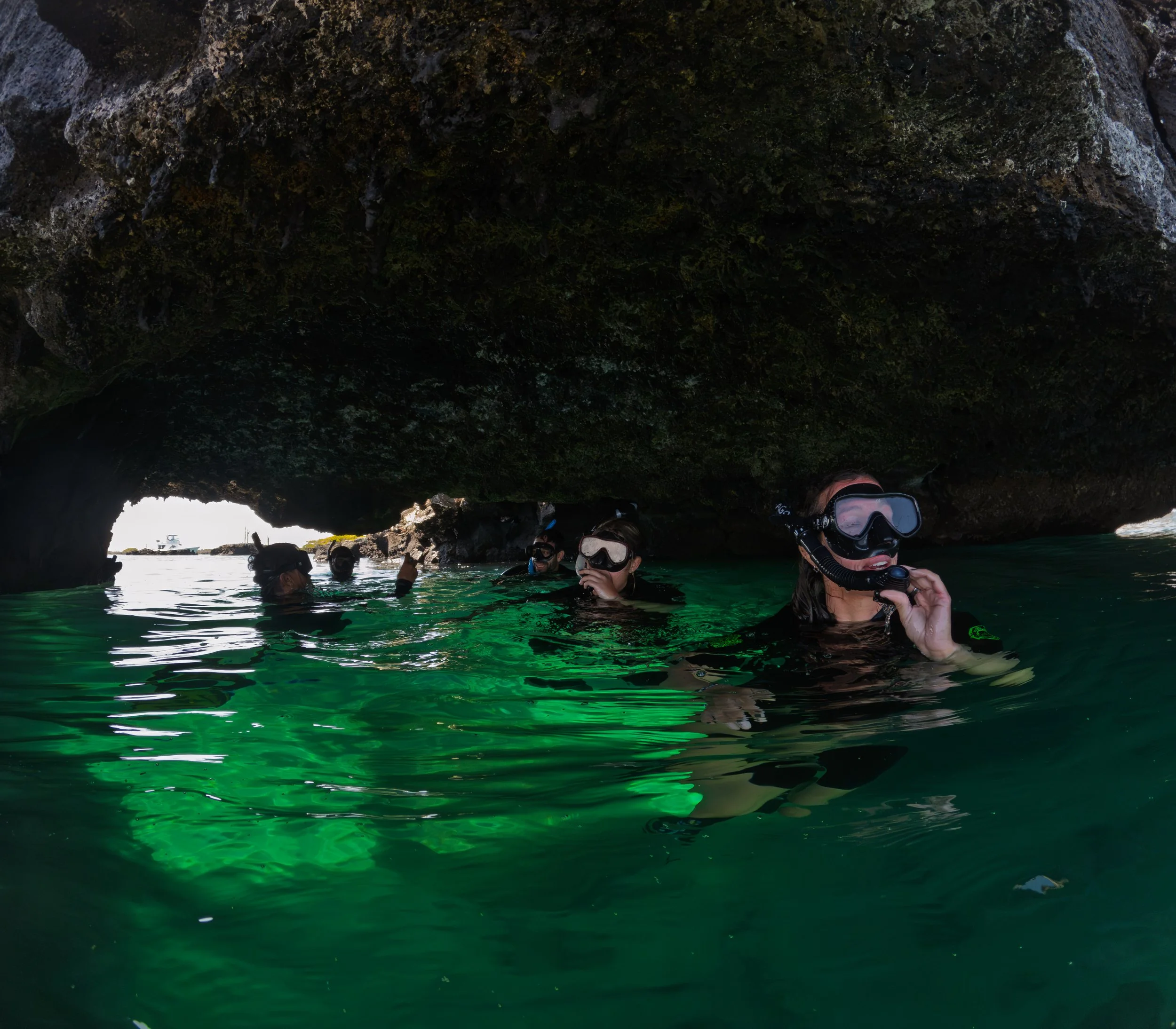 People in wetsuits snorkeling near a rocky cave with a large opening, with a boat in the water in the background.