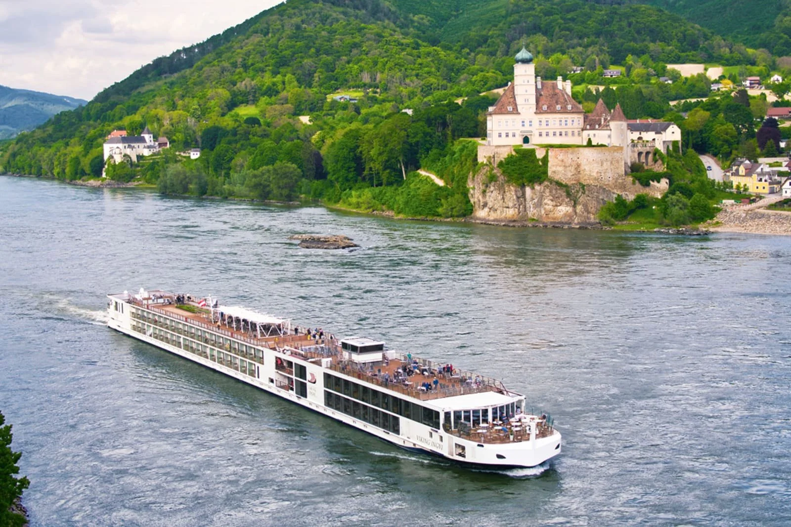 A river with a large white cruise ship sailing along, with a hill covered in green trees and a castle-like building on a rocky outcrop in the background.