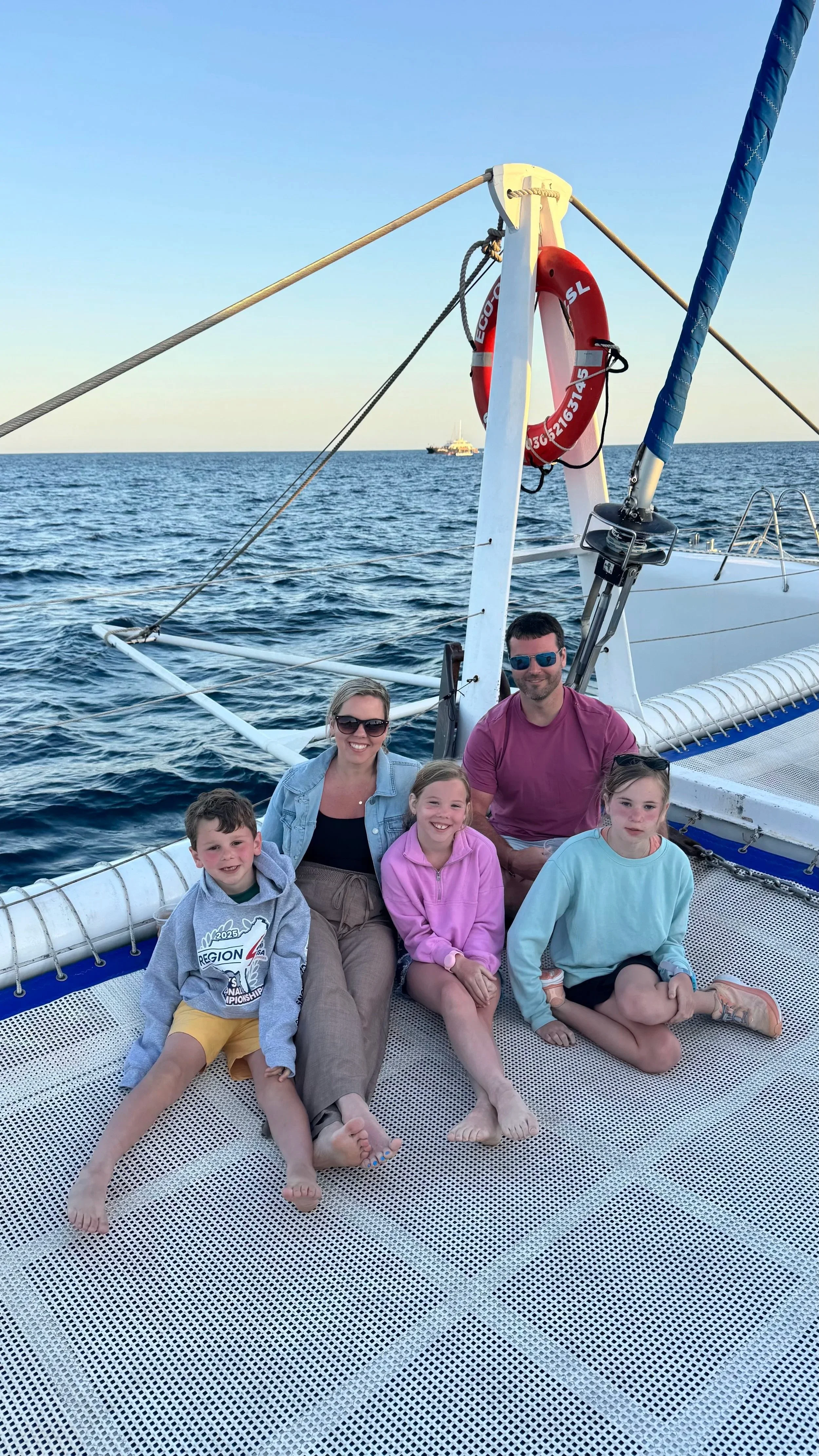 Sanders Family on a boat, sitting on a mesh deck with the ocean and a distant boat in the background, under a clear sky.