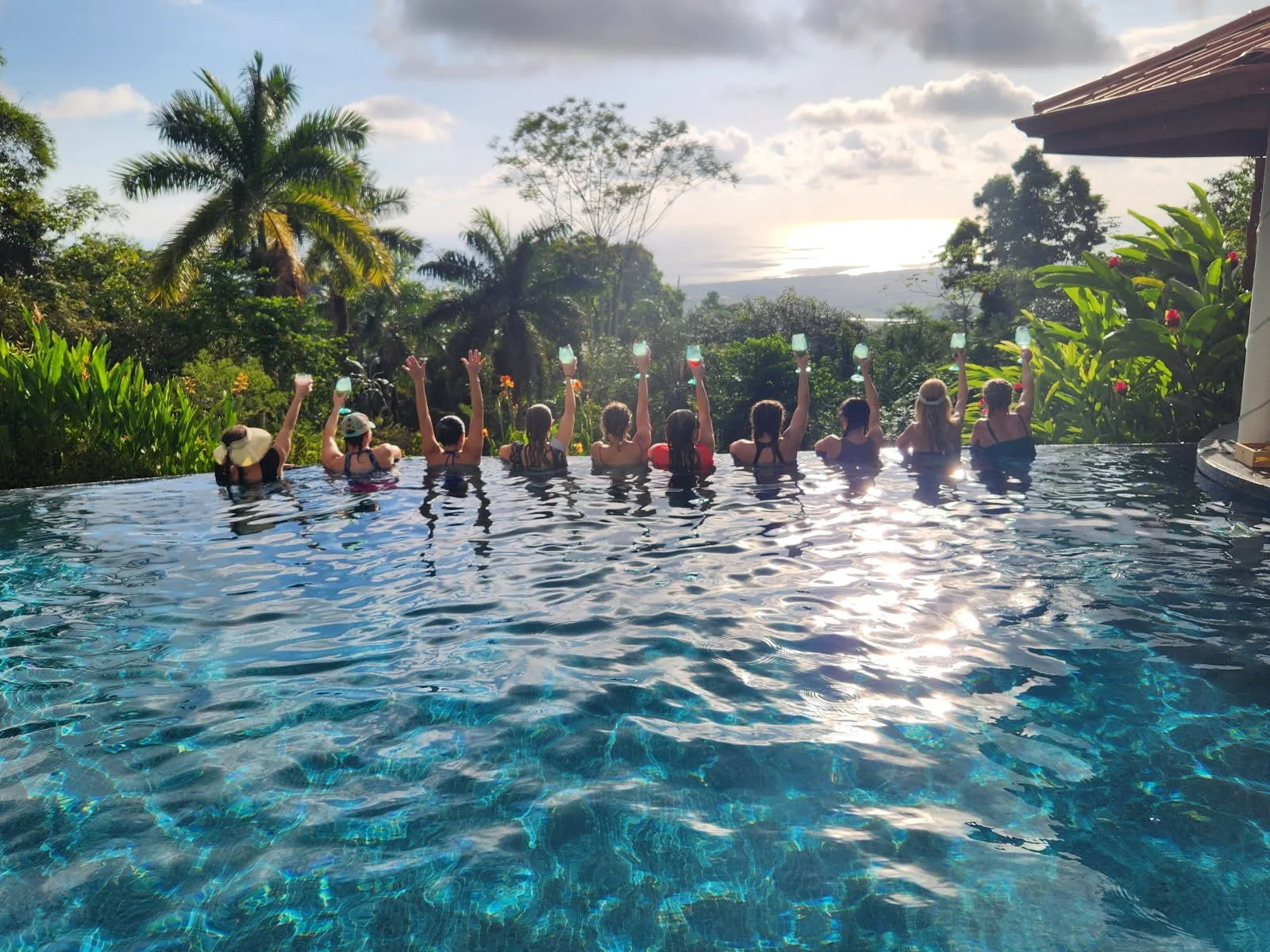 Group of people in a pool raising their glasses in a toast with a tropical landscape and sunset in the background.