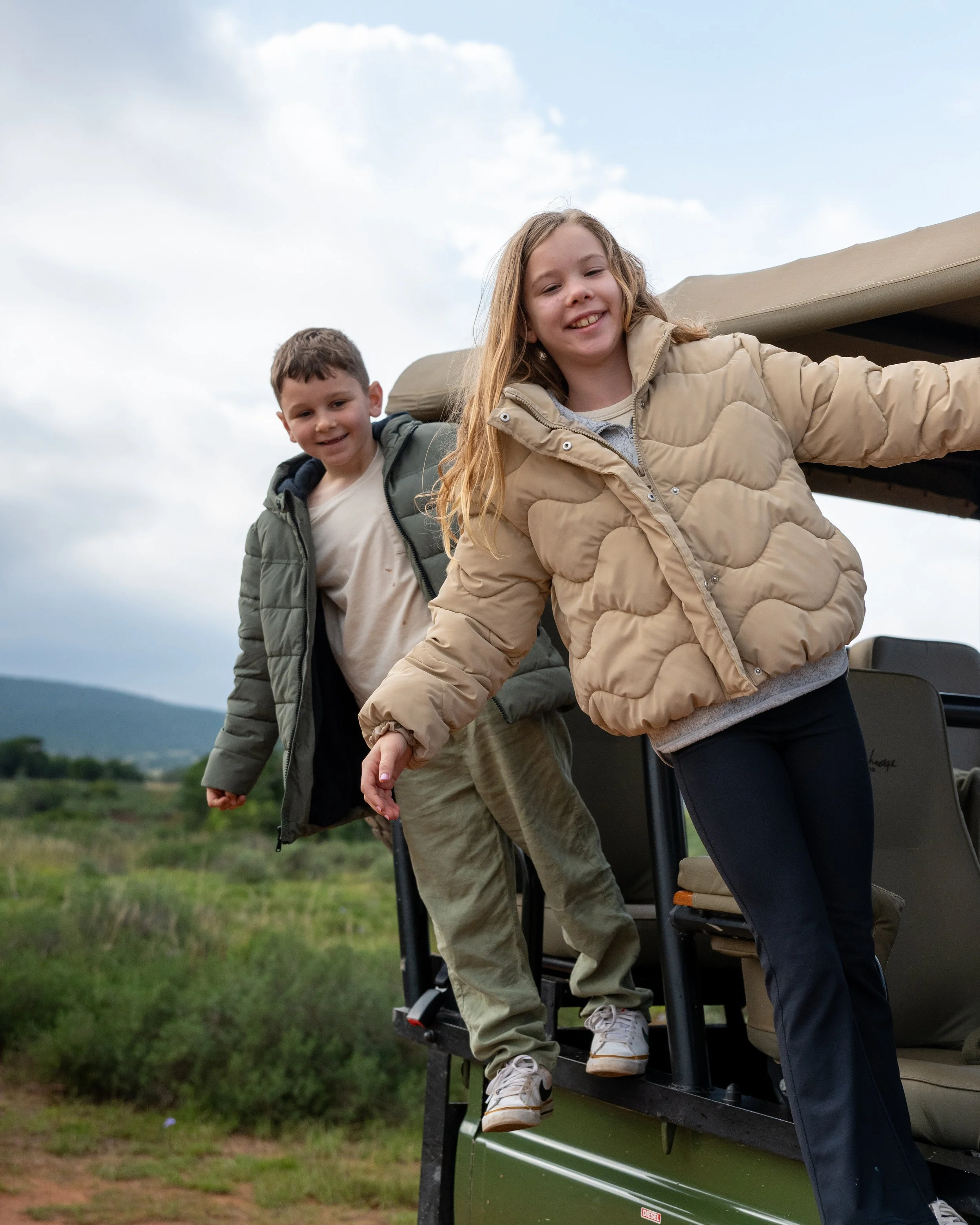 Joy & Jed, are standing outside on a grassy field, smiling, holding hands, and leaning out of an open window of a green vehicle under a cloudy sky.