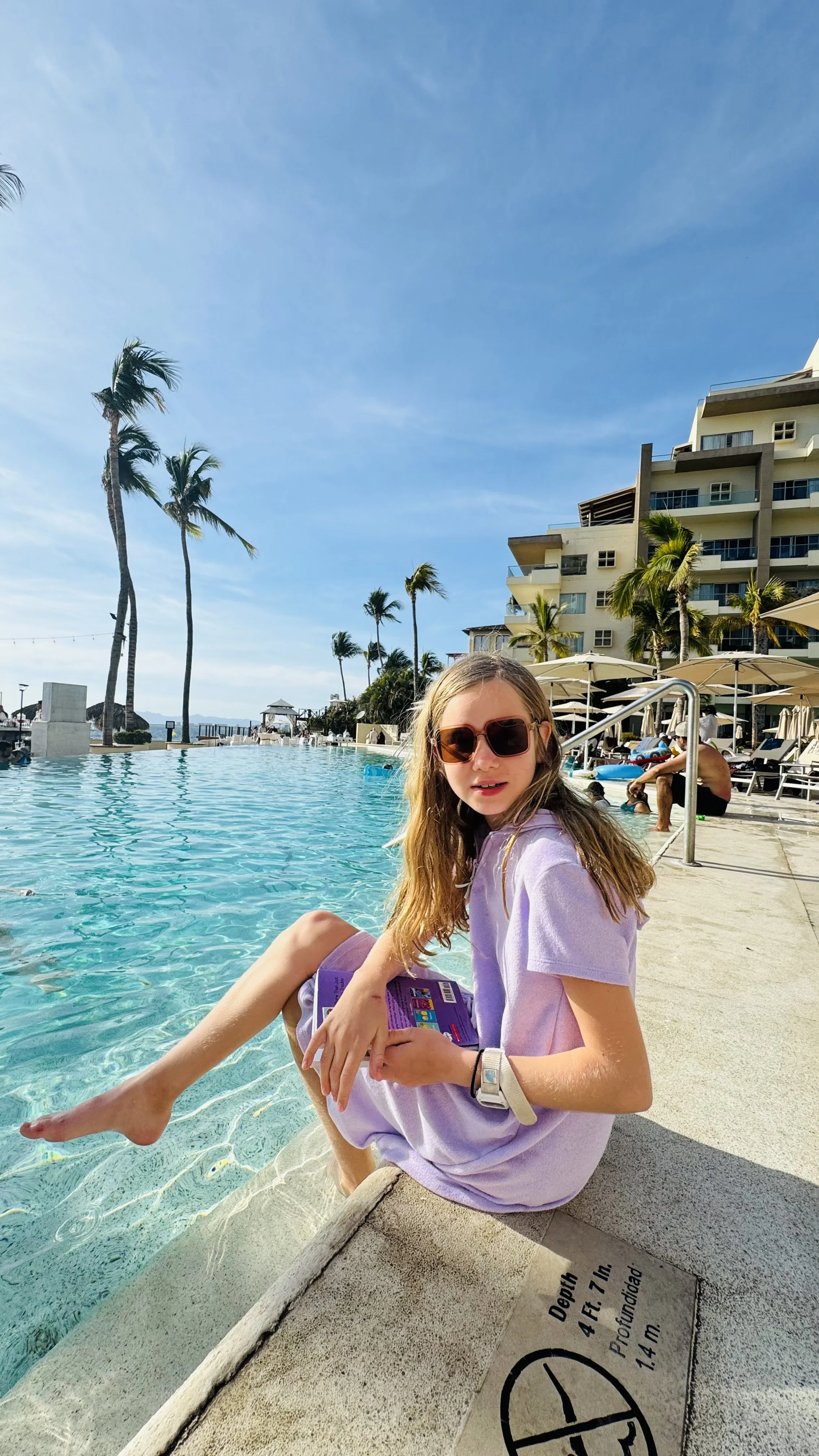 Rae wearing sunglasses and a light purple shirt sitting by a swimming pool with hotel buildings and palm trees in the background.