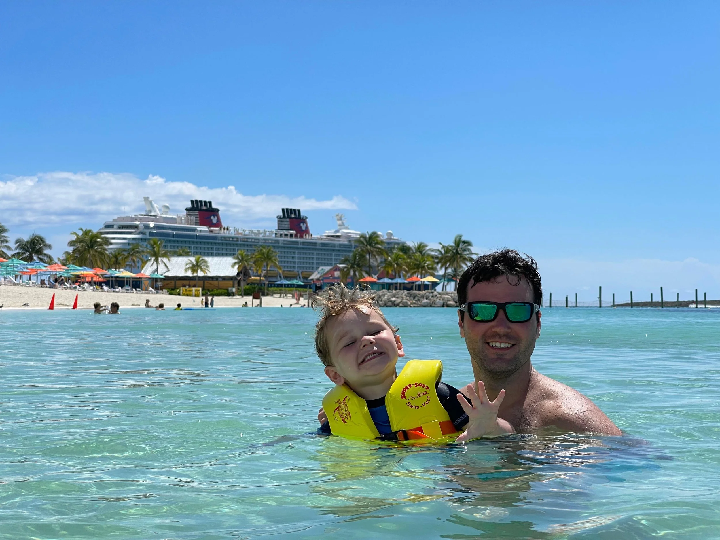 A man and a young child enjoying time in the water at a beach, with a cruise ship and blue sky in the background.