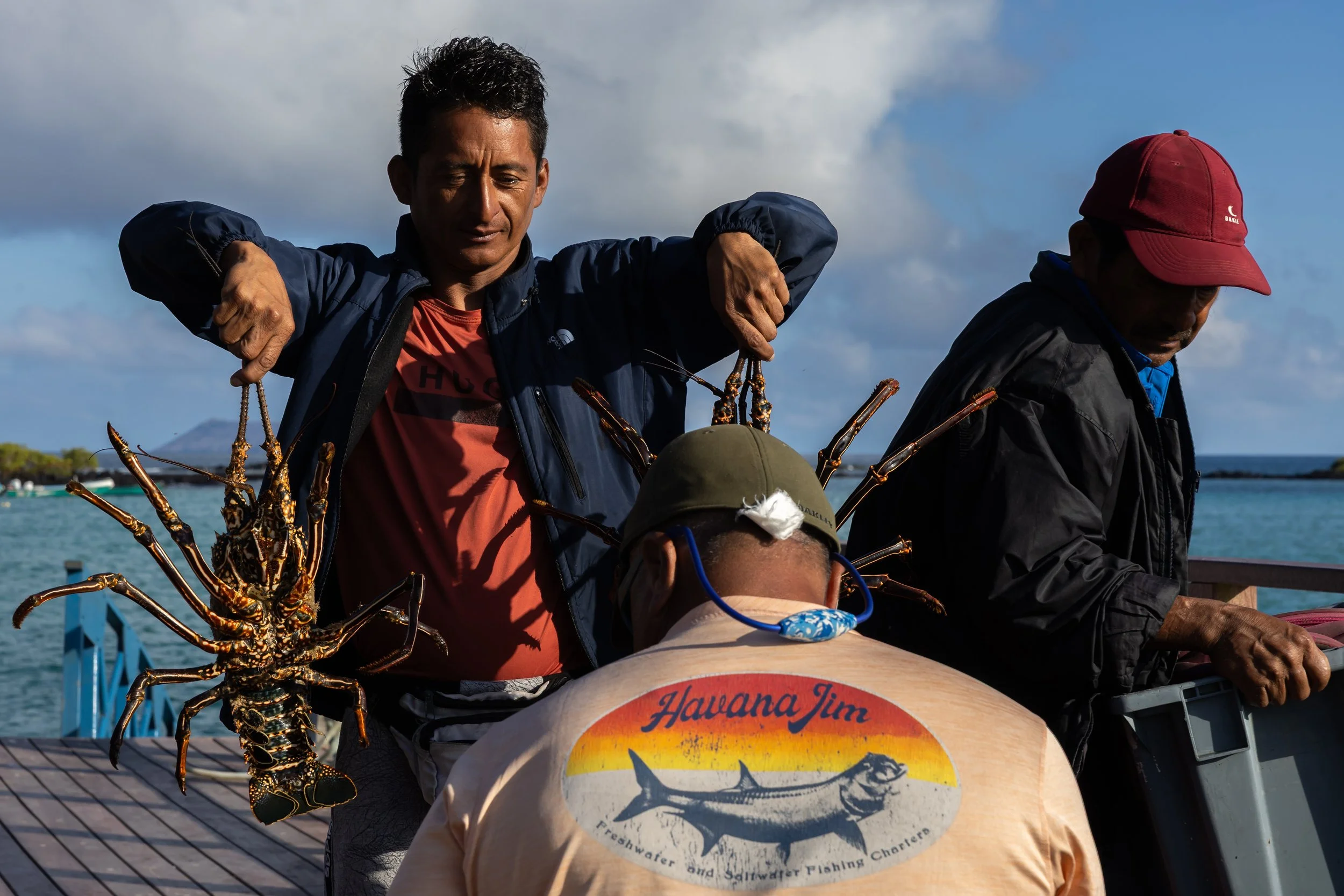 People on a boat with a large lobster in the foreground, ocean and cloudy sky in the background.
