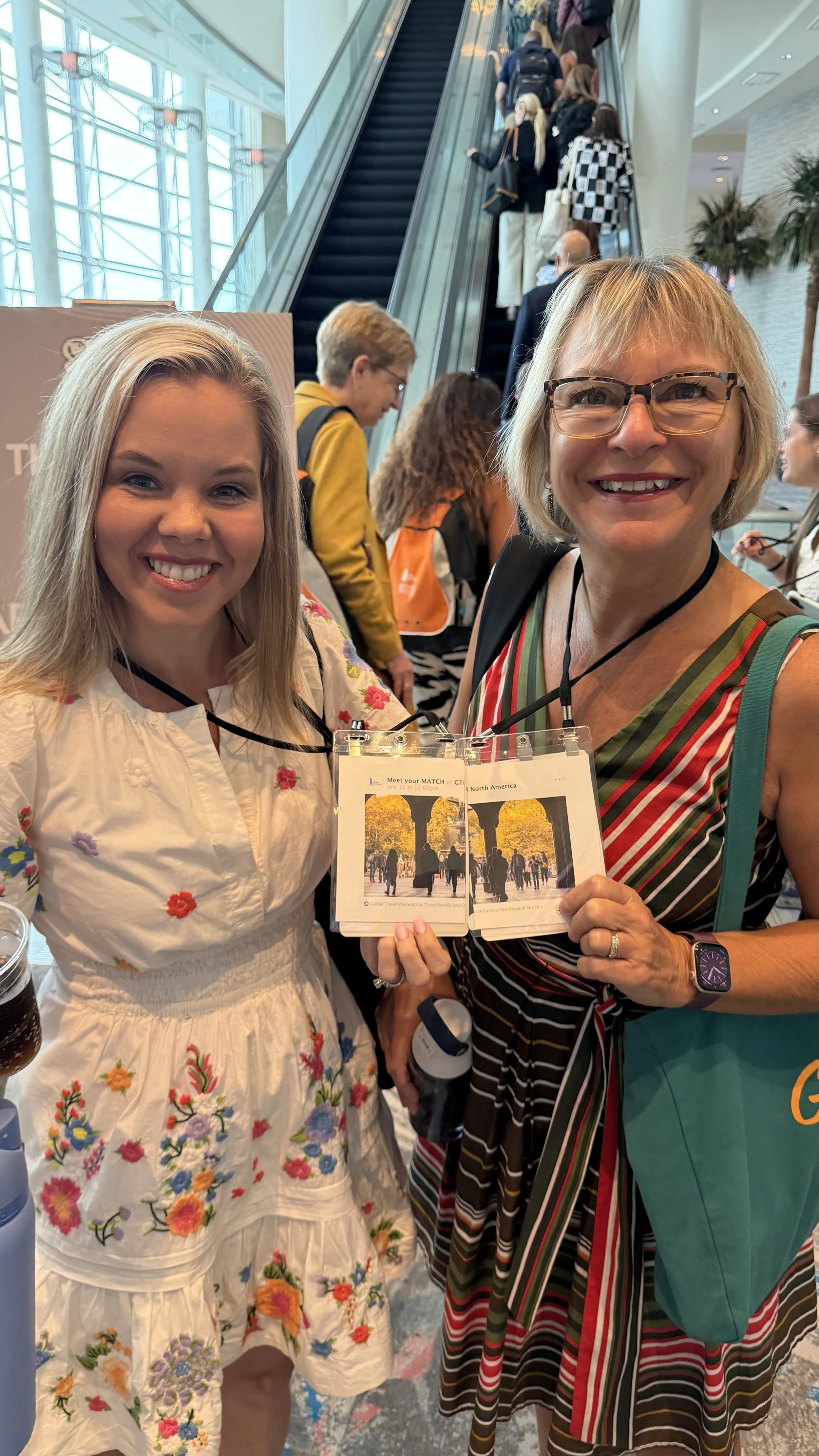 Morgan smiling and posing for a photo at an indoor event, holding various travel brochures. People are visible in the background, standing in line on an escalator in a modern, well-lit building with large windows and palm trees.