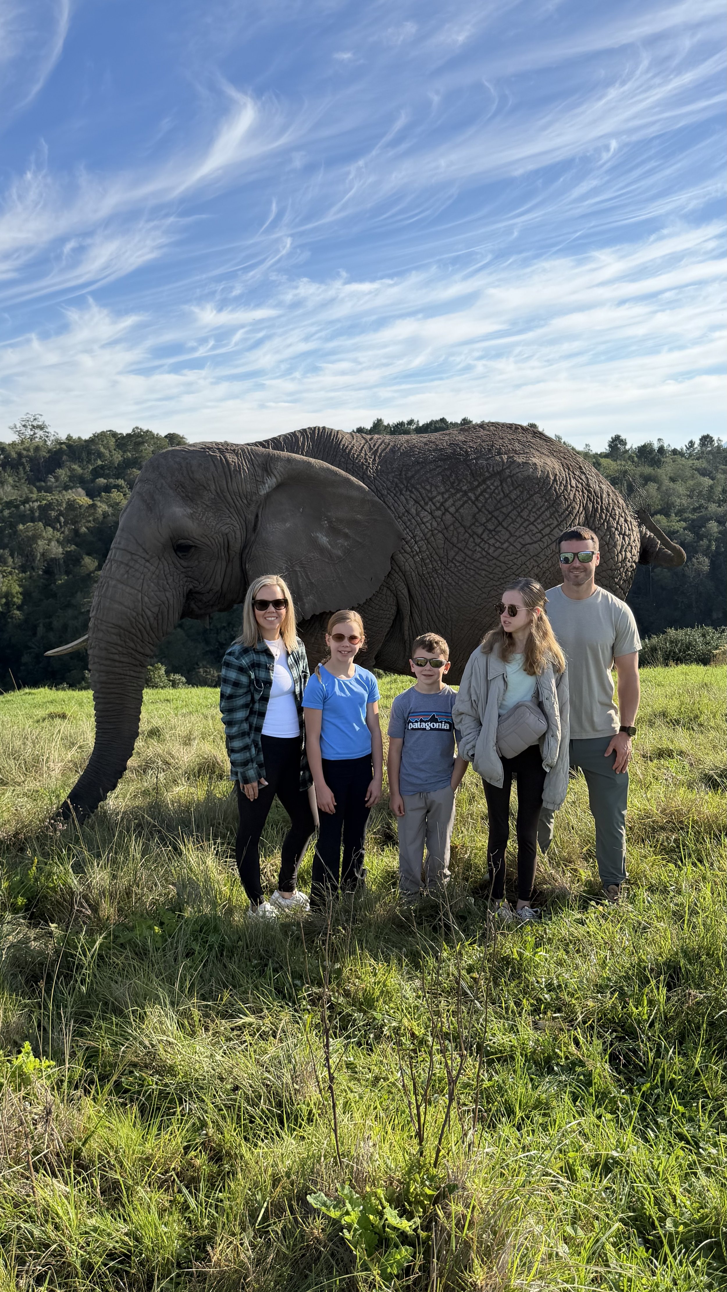 Sanders Family people standing on grass with an elephant in the background under a partly cloudy sky.
