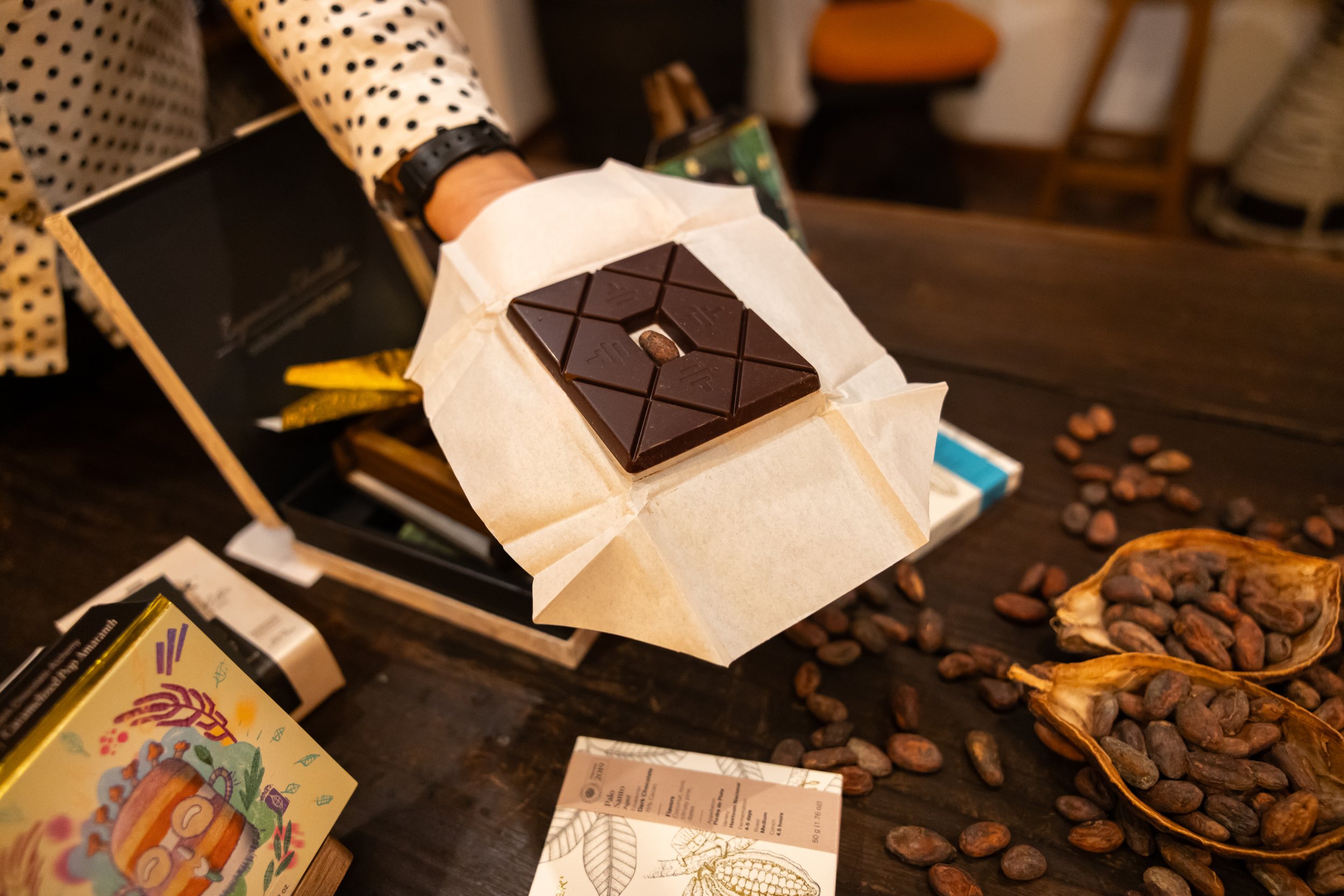 A person holding a piece of dark chocolate on parchment paper with a coffee bean in the center. The person is wearing a polka dot shirt and a black watch. There are chocolates, packaging, and coffee beans on a table.