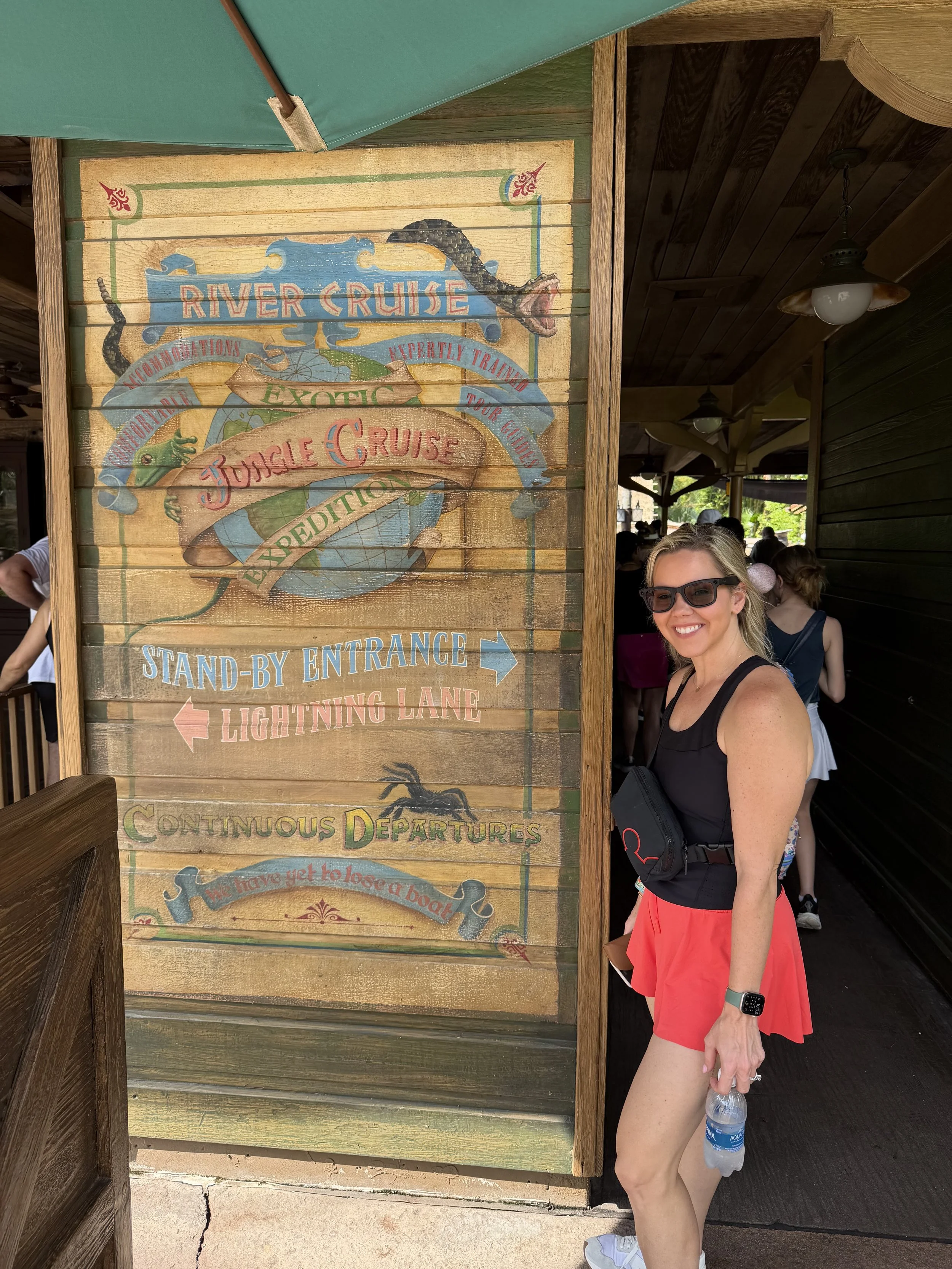 Morgan standing next to a painted sign at a theme park, smiling, holding a water bottle, wearing sunglasses, a black tank top, red shorts, and a smartwatch, with other visitors in the background.
