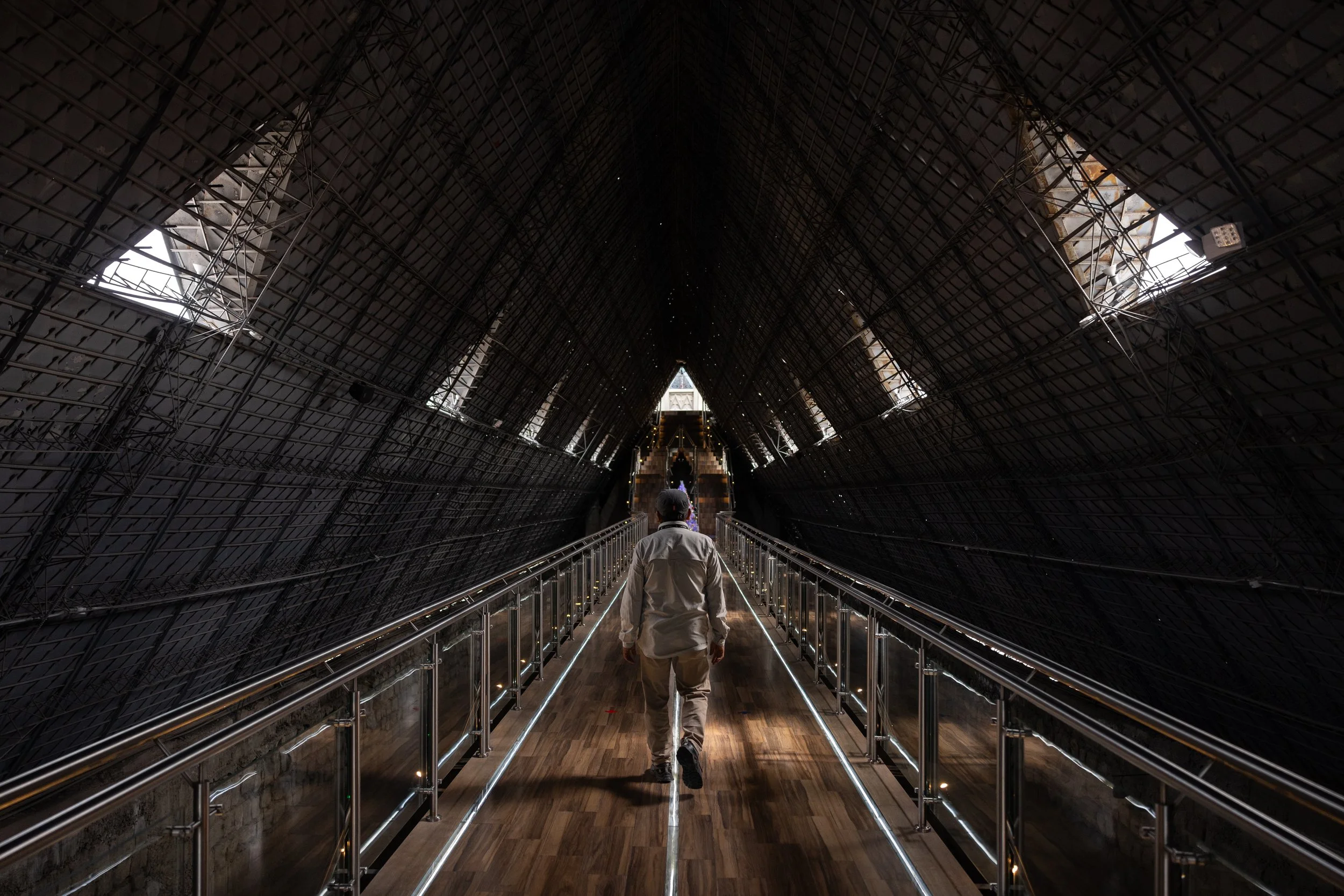 A person walking down a narrow corridor with a high, arched ceiling made of metal scaffolding, with small windows and lights before the end of the corridor.