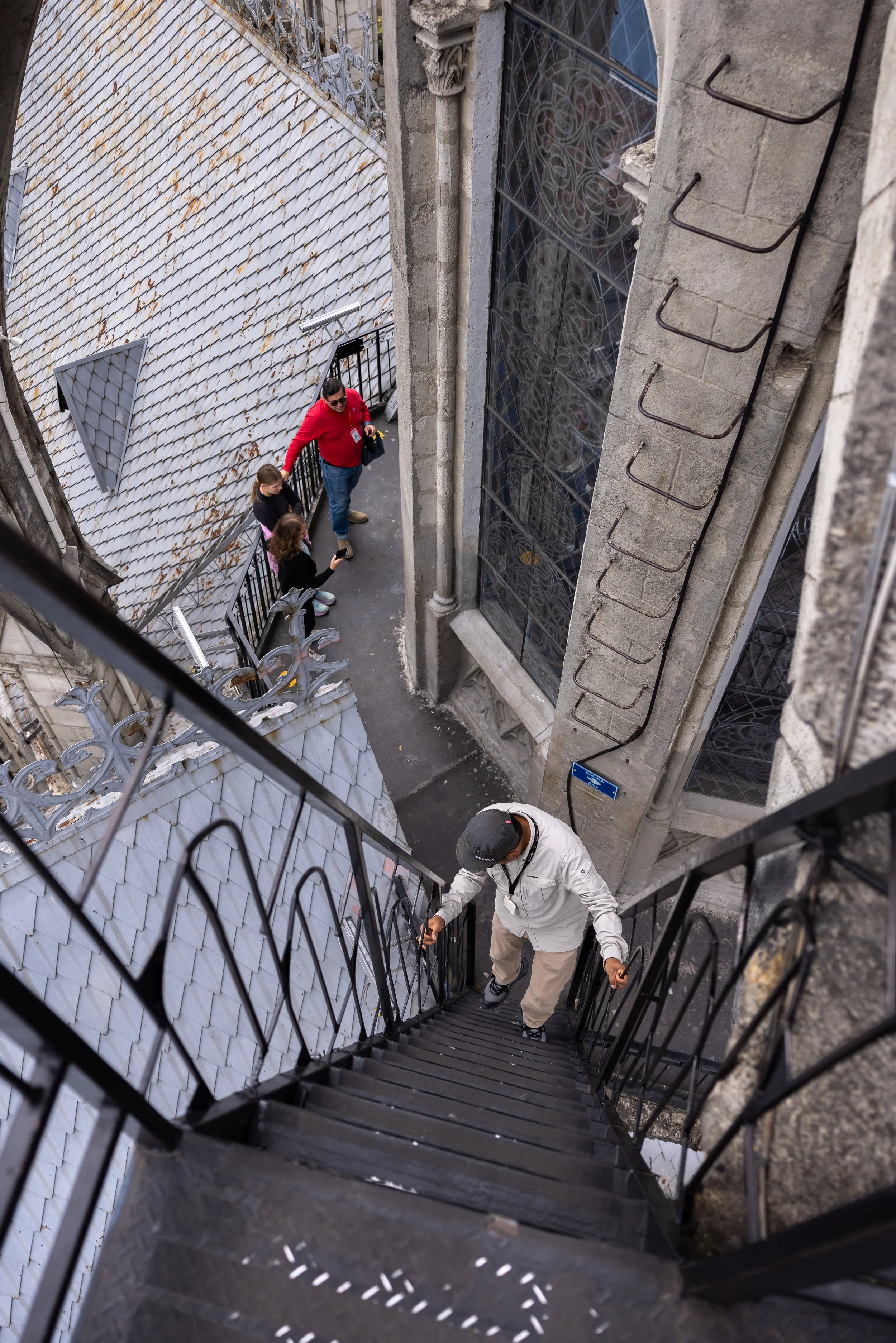 View from the top of a spiral staircase looking down, showing people climbing the stairs and architectural details of a building with stained glass windows and a tiled roof.